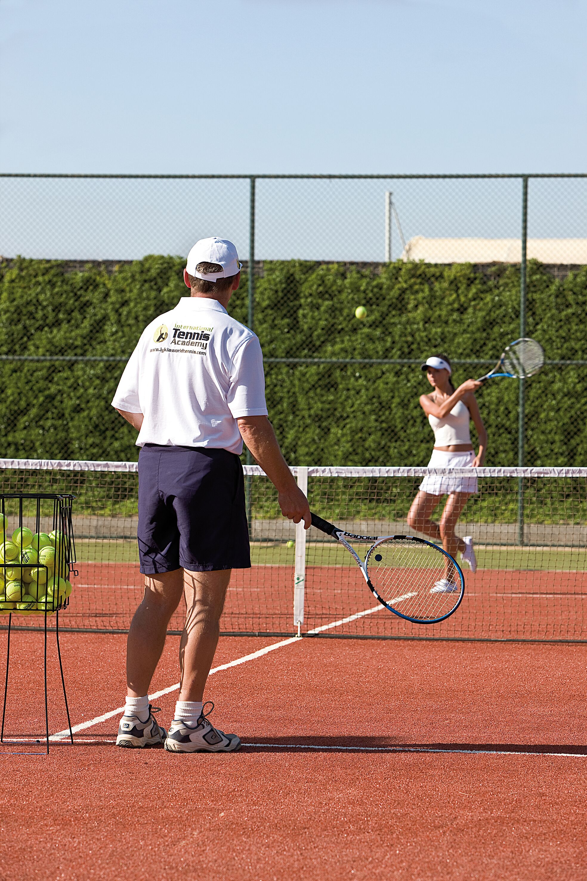Una cancha de tenis con una red, una máquina de pelotas y un portaobjetos para pelotas.