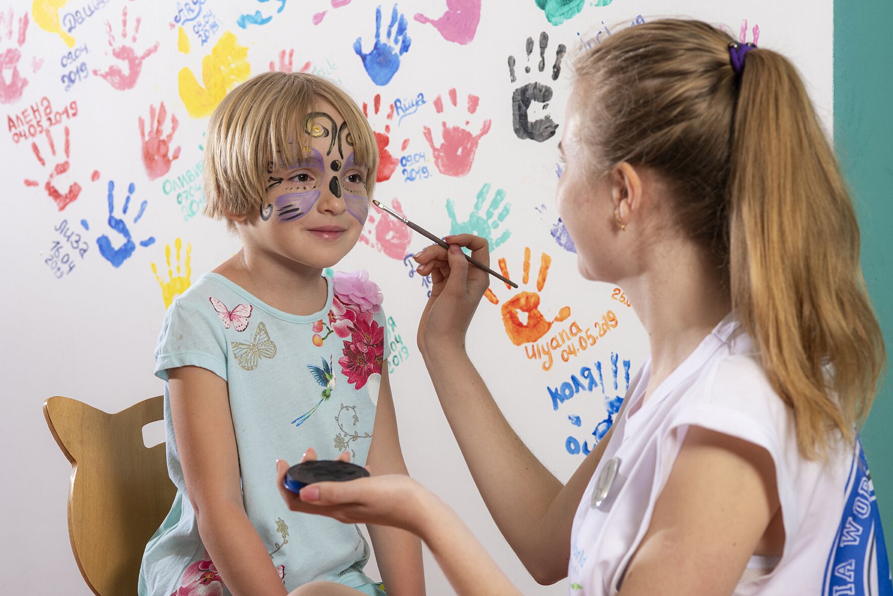 Niño recibiendo una sesión de pintura facial en una habitación con huellas de manos coloridas en la pared.