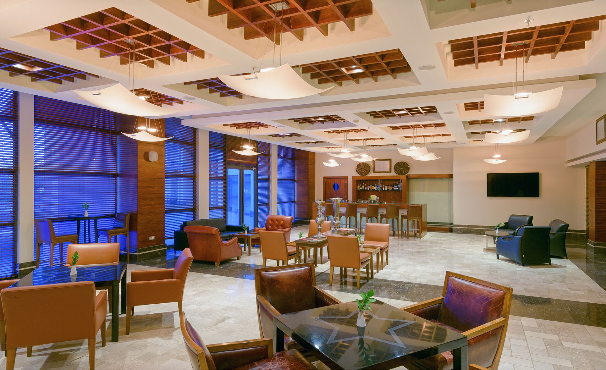 Un vestíbulo amplio con mobiliario moderno, una zona de bar y ventanas grandes con persianas.