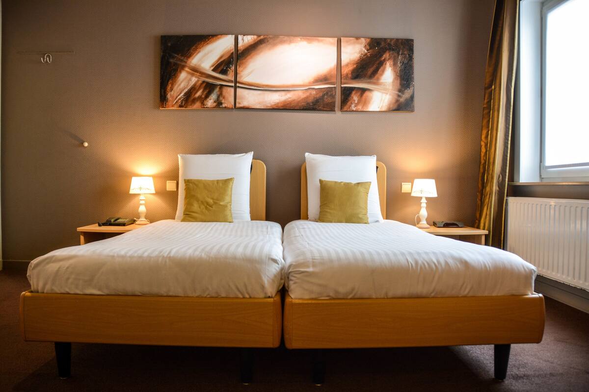 A hotel room with two beds, each with a pillow and a bedside lamp. There is a large abstract painting on the wall above the beds.
