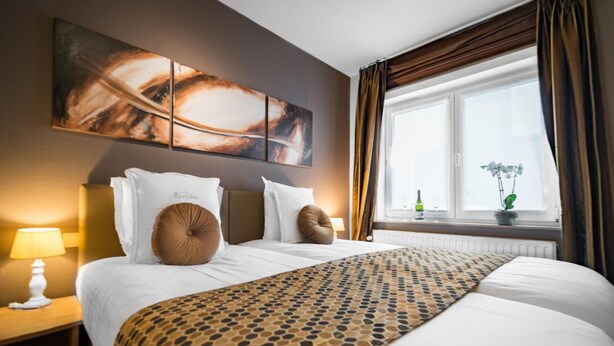 A hotel room with a large bed, a nightstand with a lamp, a window with curtains, and a wall with abstract art. - Hotel Maraboe (Bruges)