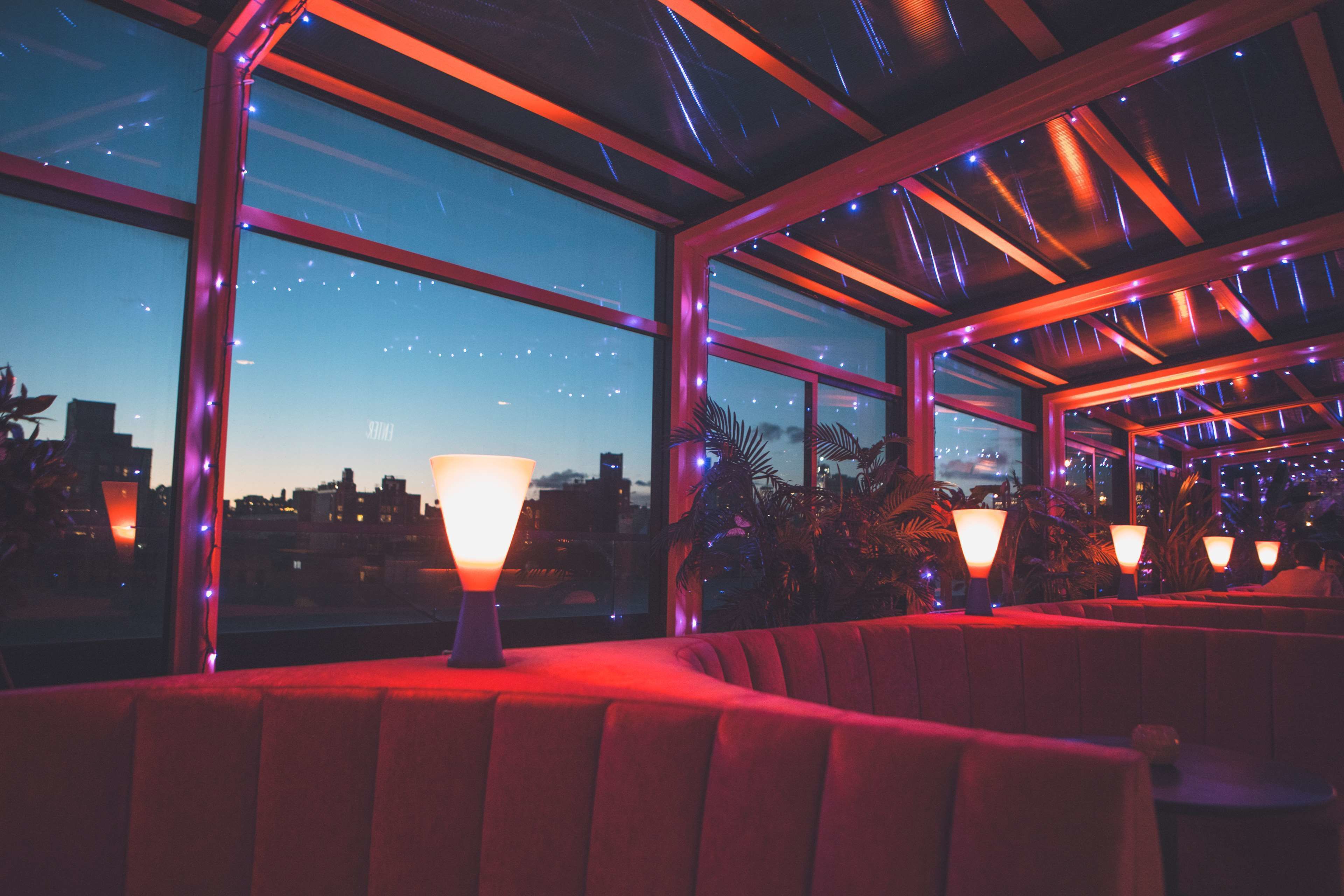 A rooftop bar with red seating, large windows, and city views at dusk.
