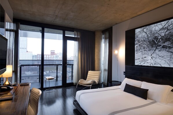 A hotel room with a large bed, a desk with a lamp, a chair, a small table, and a view of the city through large windows.