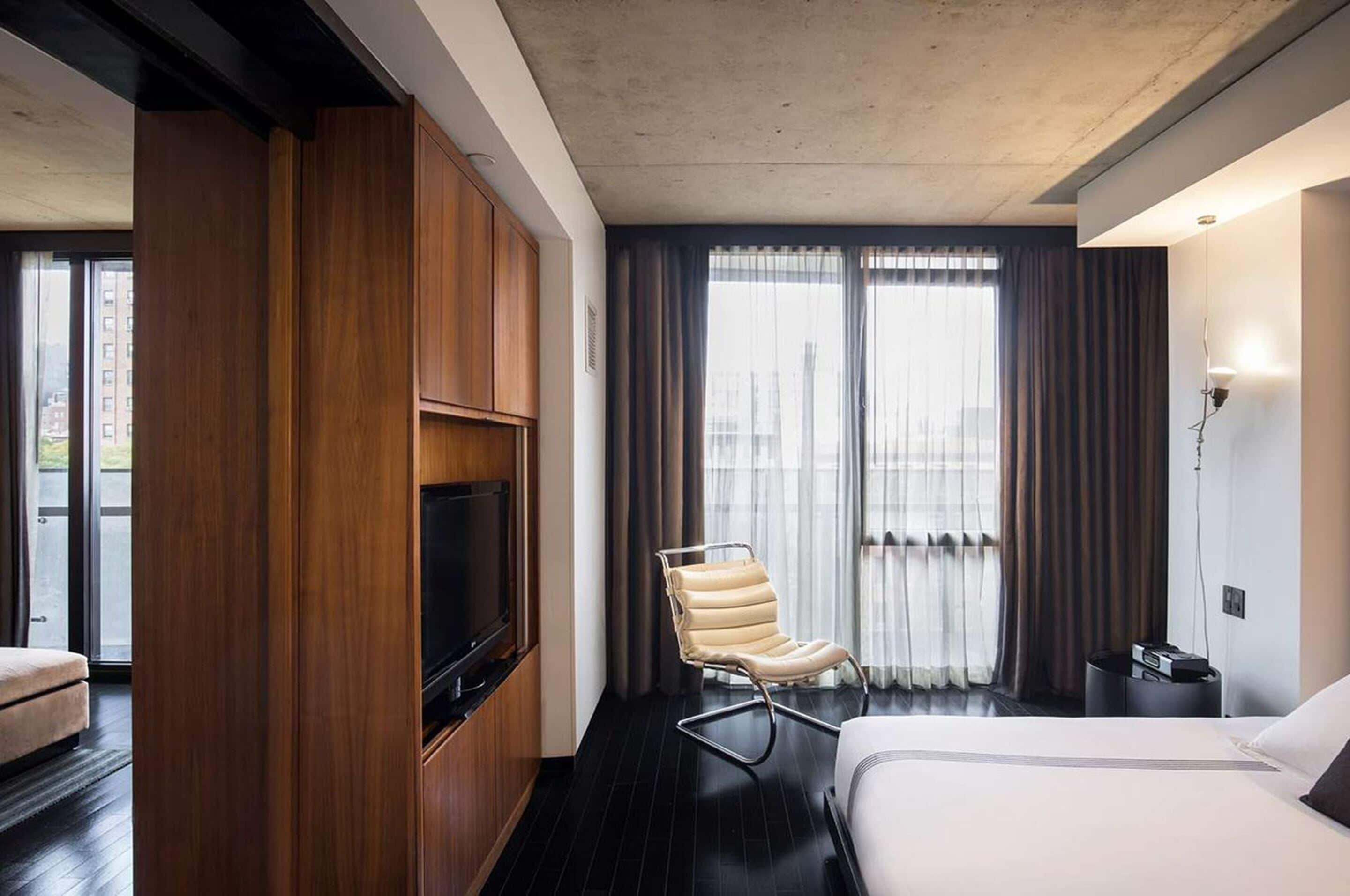 Una habitación de hotel con cama, televisión, armario de madera, una silla y ventana con cortinas.
