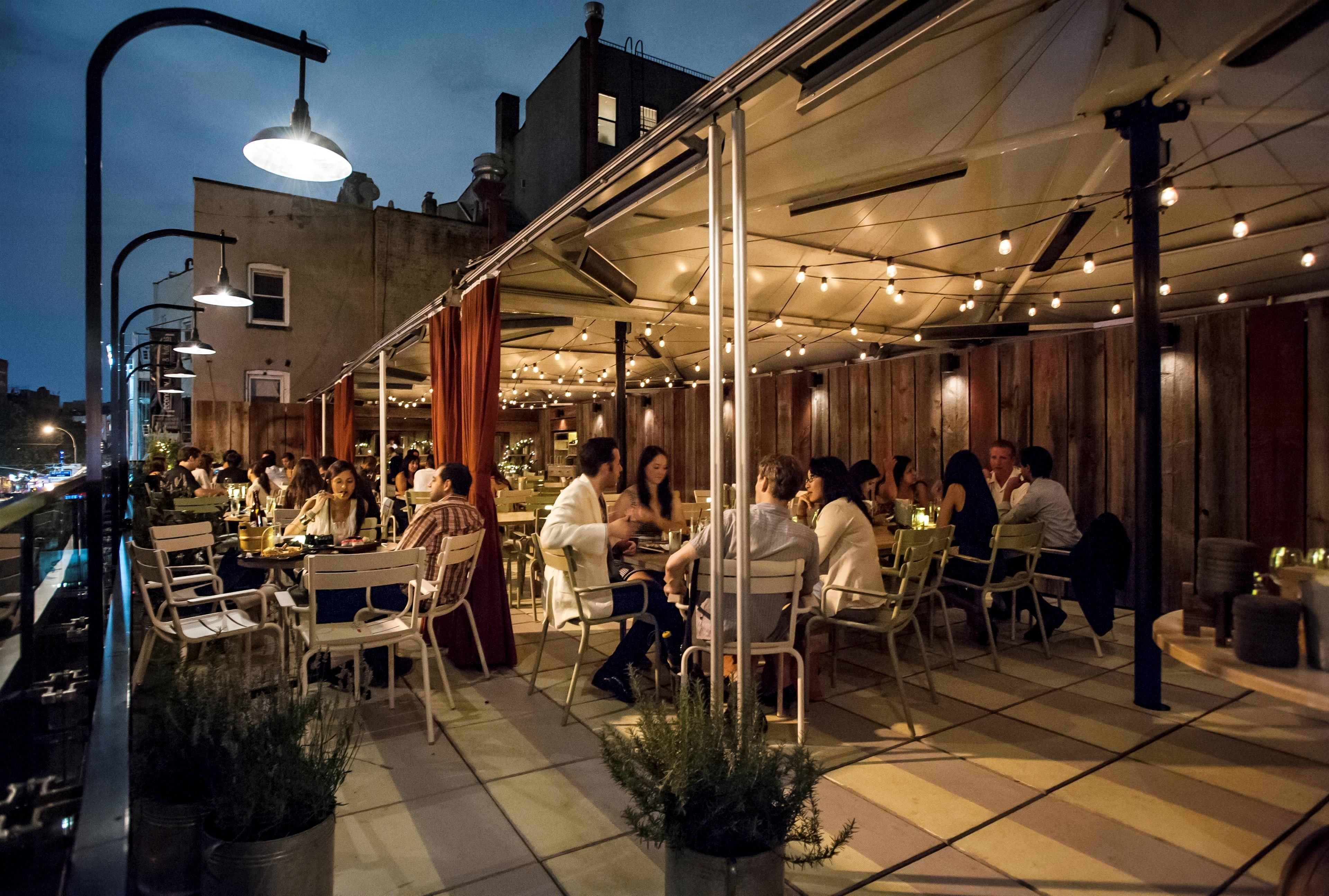 An outdoor dining area with tables and chairs, a covered section with string lights, and a view of the cityscape in the background.