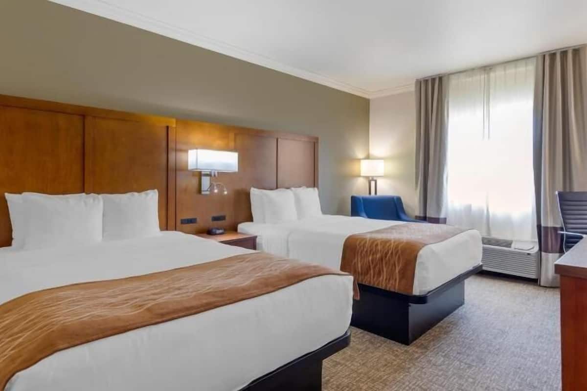 A hotel room with two beds, a wooden headboard, a desk, a chair, and a window with curtains.