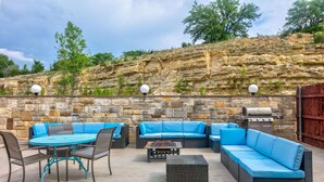 Une terrasse extérieure avec un mur en pierre, des sièges rembourrés bleus, une table ronde et un barbecue.