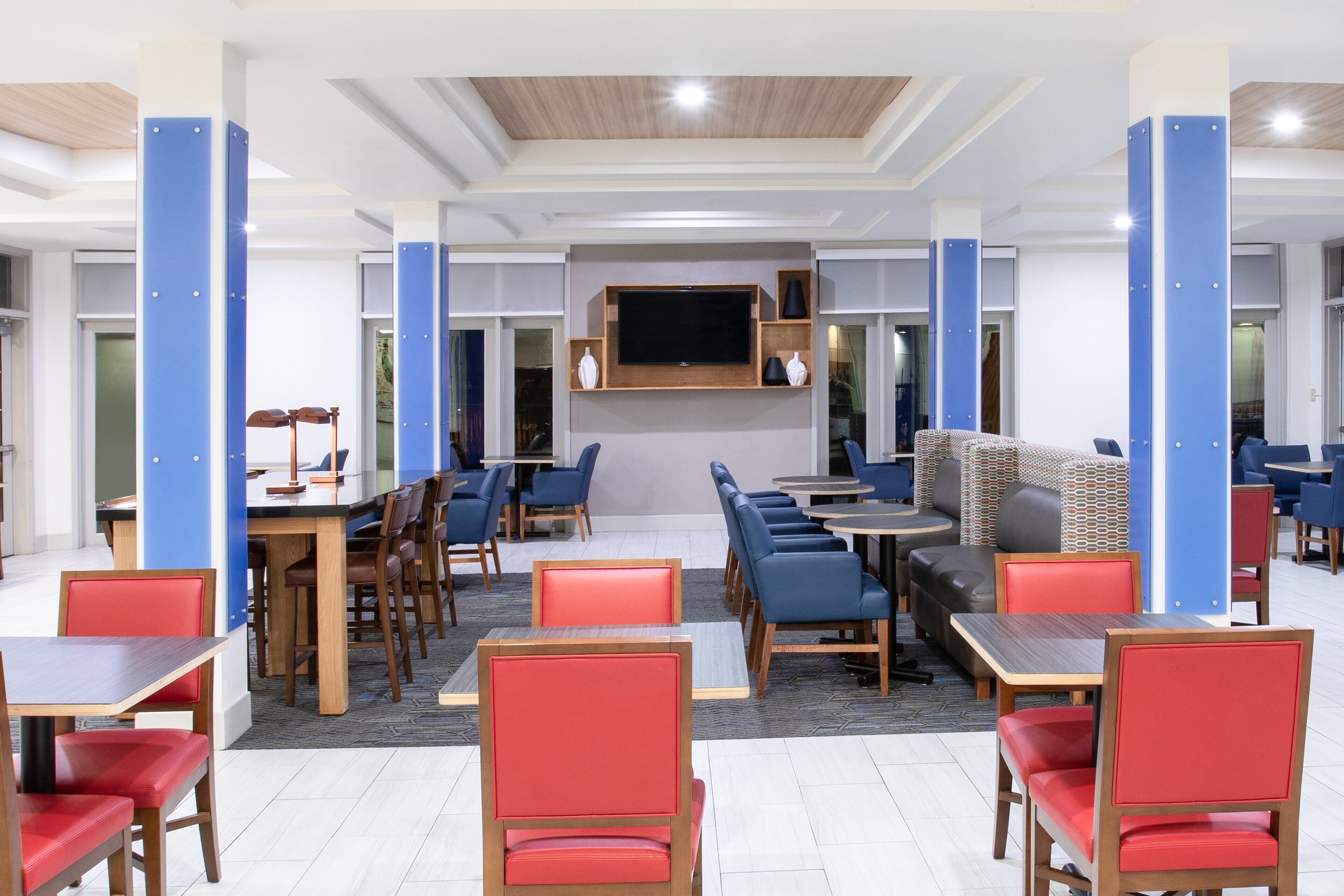 A hotel lobby with multiple seating areas, a large flat-screen TV mounted on the wall, and a variety of tables and chairs.
