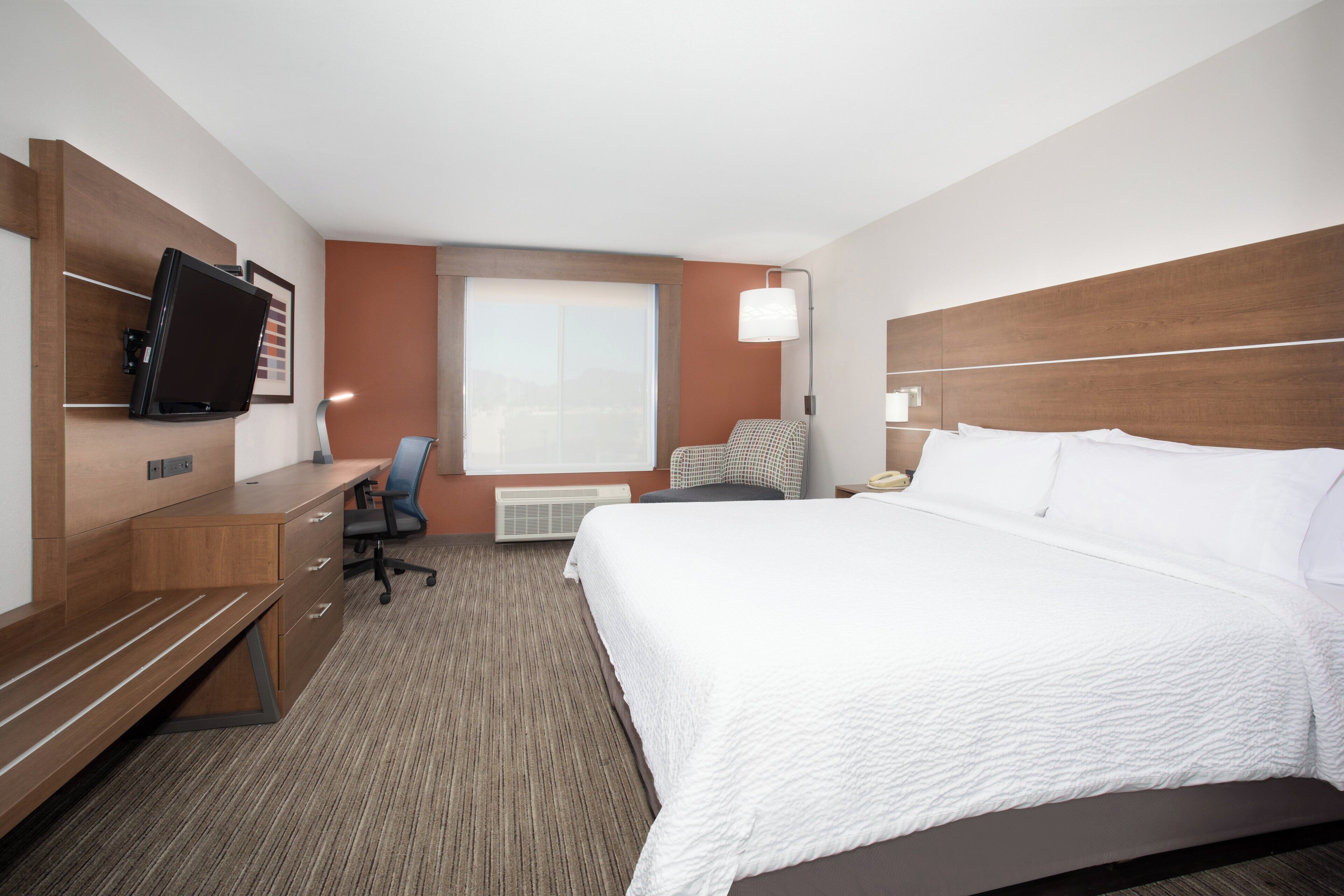 A hotel room with a bed, a desk with a chair, a TV, a window, and a lamp.
