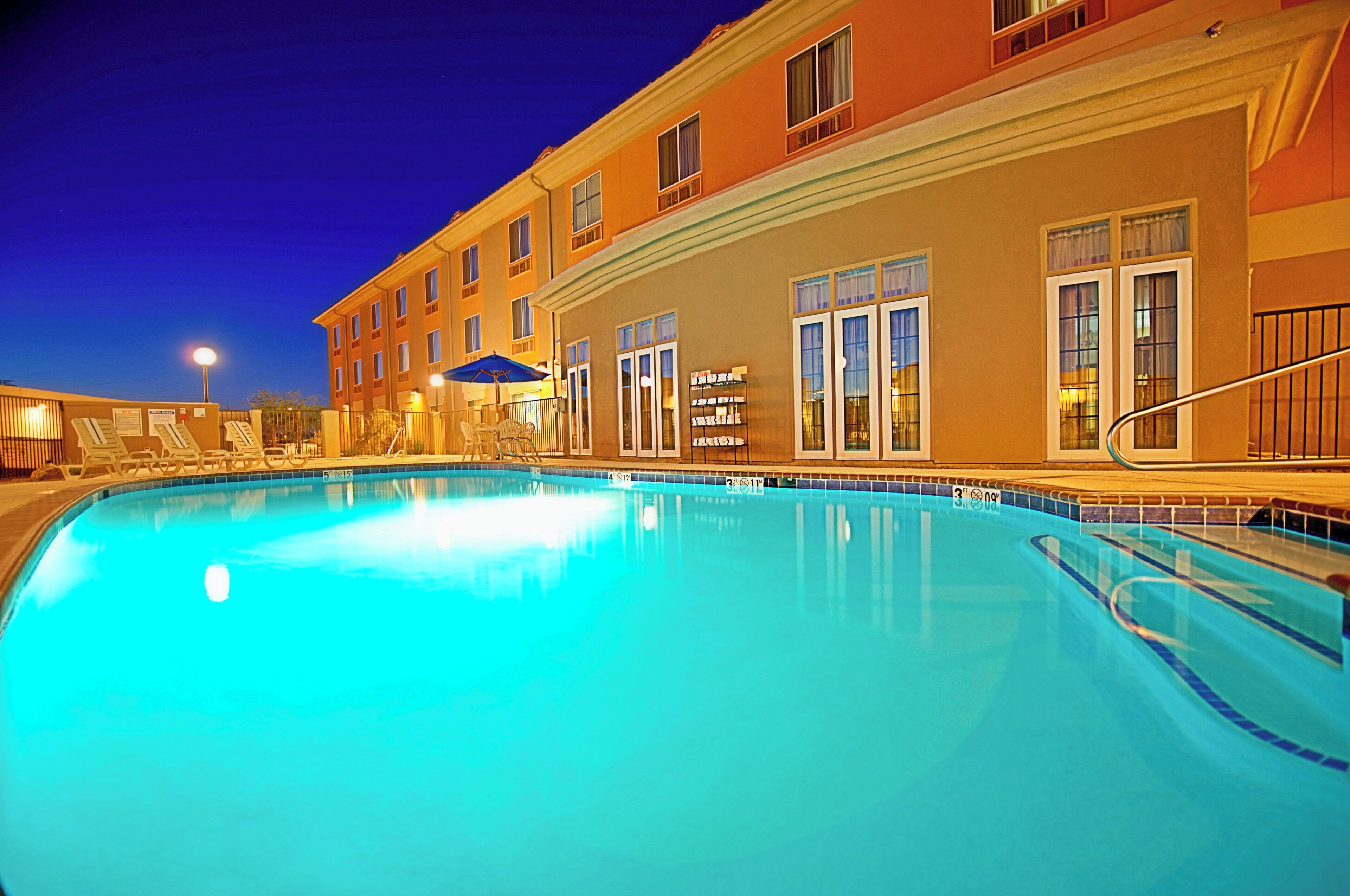 A swimming pool with lounge chairs and umbrellas, surrounded by a building with multiple windows and a balcony.