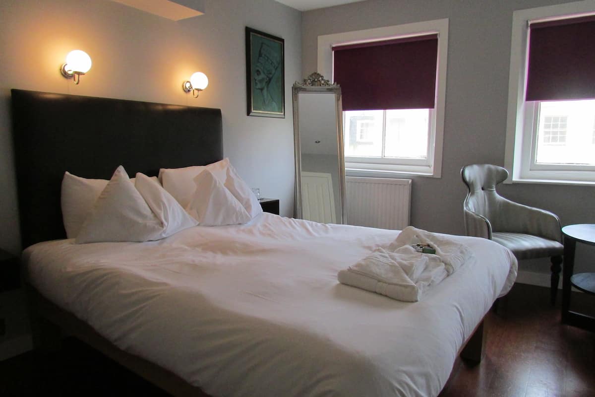Een net opgemaakt bed met witte lakens en kussens, een stoel, een spiegel en twee wandlampen.
