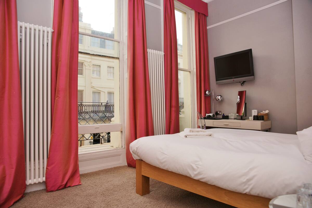 Hotelkamer met een bed, een aan de muur bevestigde televisie, een raam met rode gordijnen en uitzicht op gebouwen.
