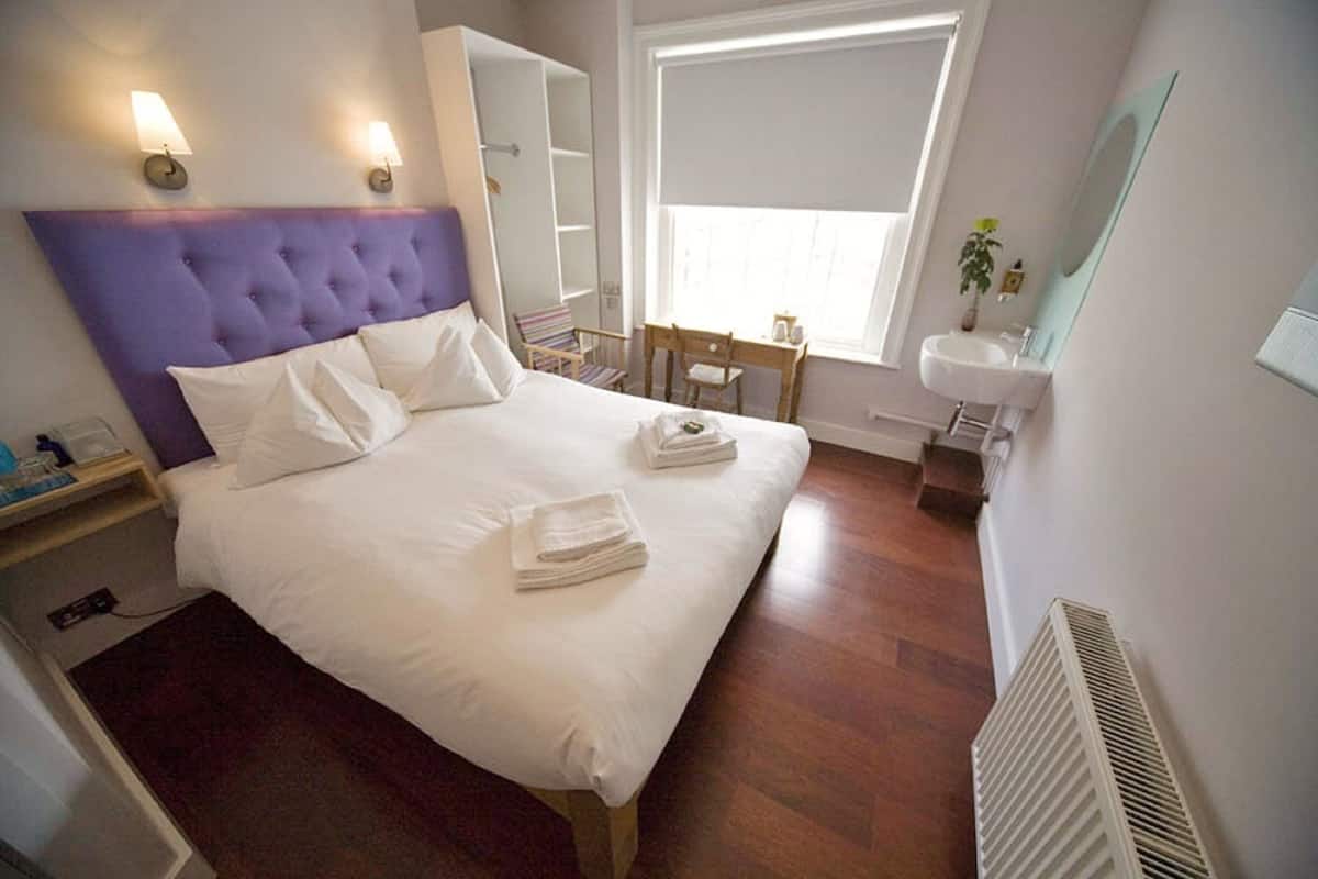 Een net opgemaakt bed met witte lakens en handdoeken, een nachtkastje met een lamp, een raam met zonwering, een klein bureau met een stoel, en een witte radiator.