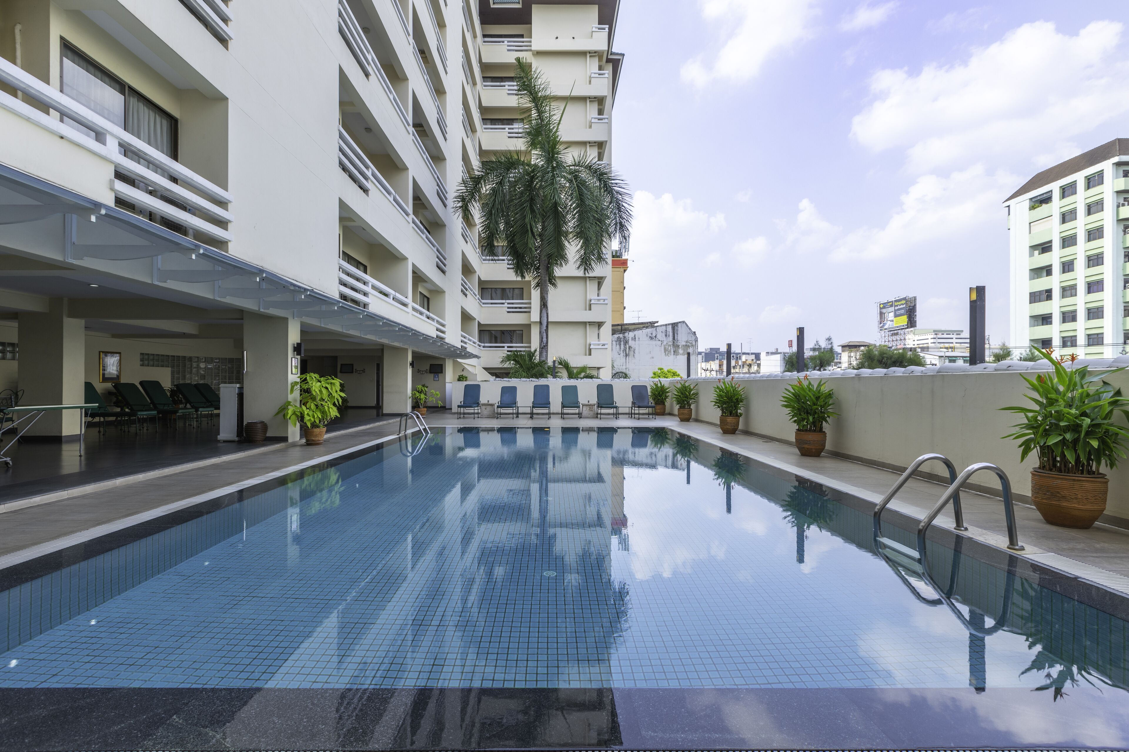 A swimming pool with a clear blue tile surface, surrounded by a white building with balconies and potted plants.