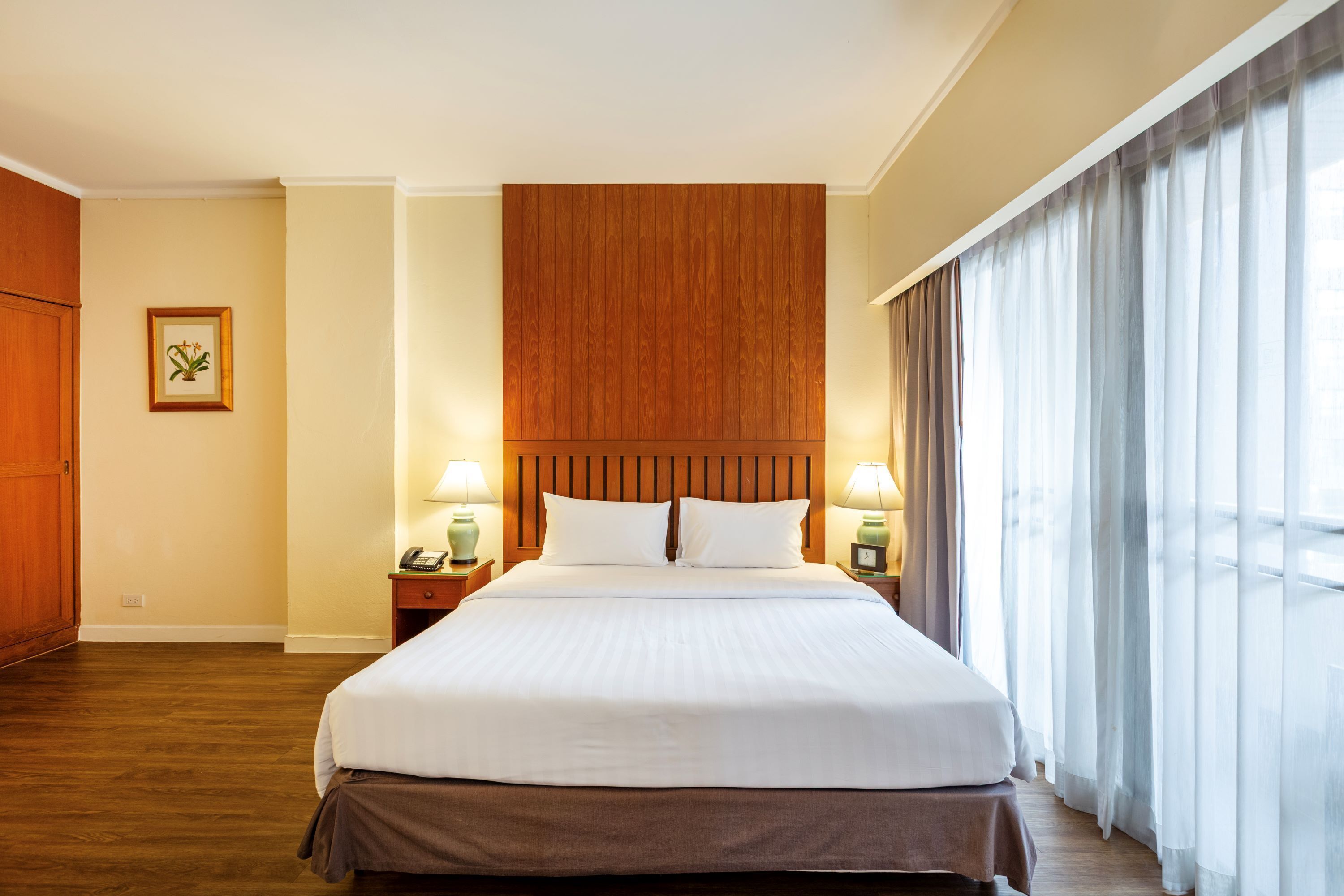 A hotel room with a large bed, wooden headboard, two bedside lamps, a nightstand, and a window with sheer curtains.