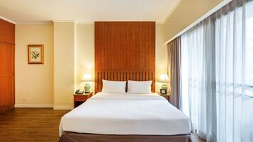 A hotel room with a large bed, wooden headboard, two bedside lamps, a nightstand, and a window with sheer curtains.
