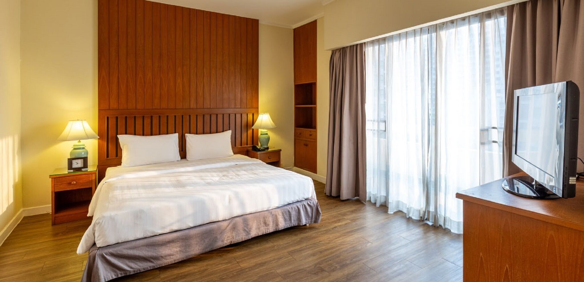A hotel room with a large bed, wooden headboard, two bedside tables with lamps, a TV on a wooden stand, and a window with curtains.