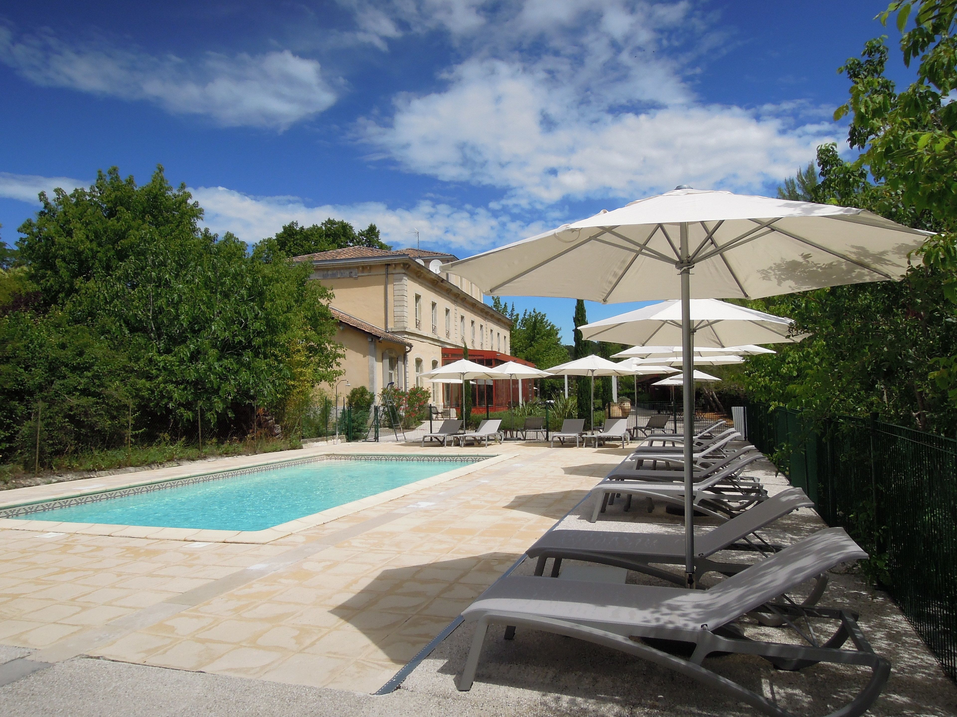 Un espace piscine avec des chaises longues et des parasols, entouré de verdure et avec un bâtiment en arrière-plan.
