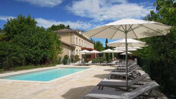 Un espace piscine avec des chaises longues et des parasols, entouré de verdure et avec un bâtiment en arrière-plan.