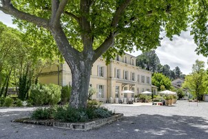 Um pátio com uma grande árvore, mesas ao ar livre e um edifício com várias janelas.
