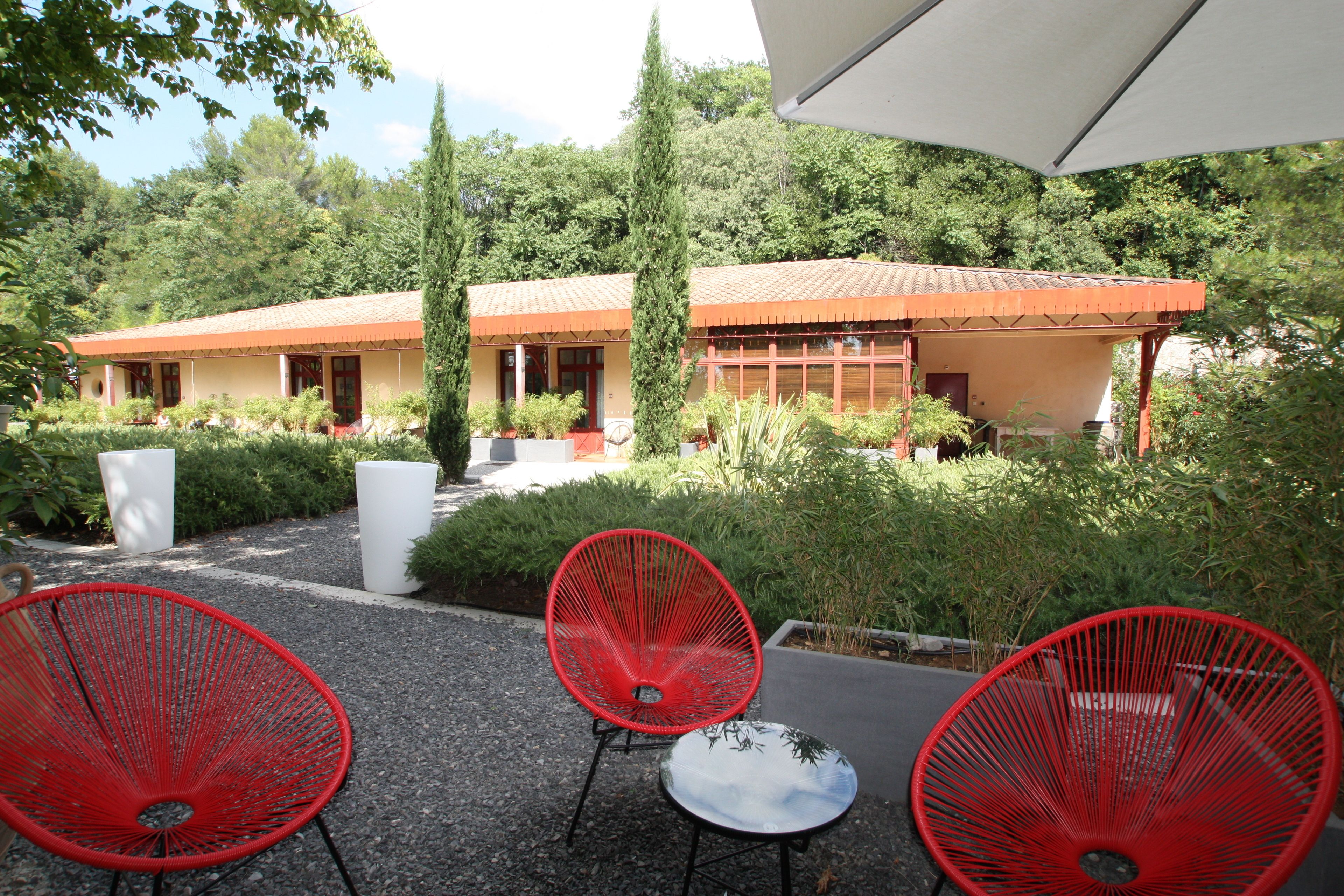 Un patio con sillas rojas y una mesa, rodeado de follaje y un edificio con tejado de baldosas.