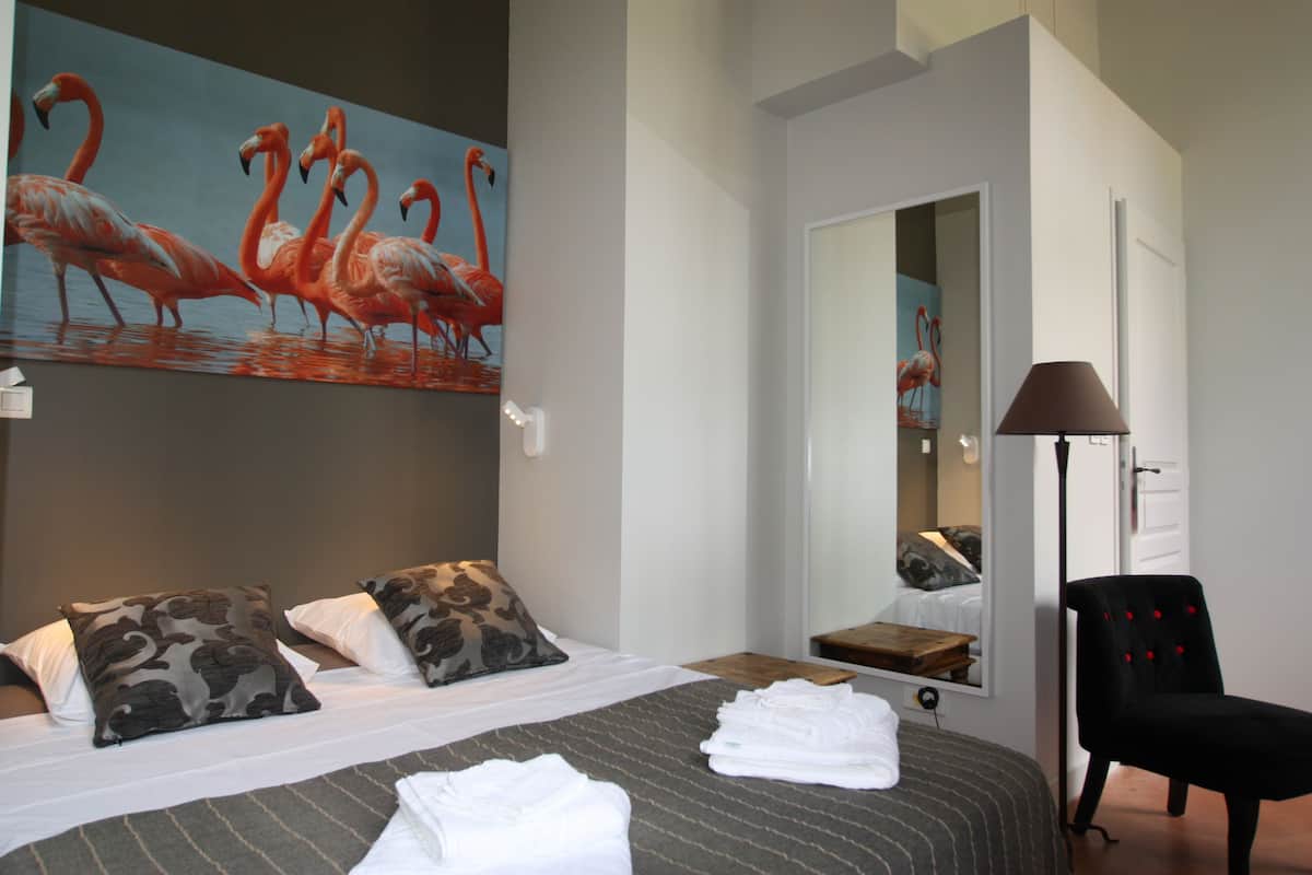 A hotel room with a bed, pillows, a painting of flamingos, a chair, and a mirror.