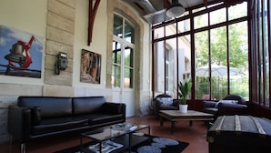 A spacious living room with a black leather sofa, a glass coffee table, and a vintage suitcase. There are large windows offering a view of greenery outside. - Hotel Estelou (Sommieres)