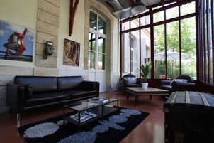 Uma sala de estar espaçosa com um sofá de couro preto, uma mesa de centro de vidro e uma mala antiga. Janelas grandes oferecem vista para o verde lá fora.