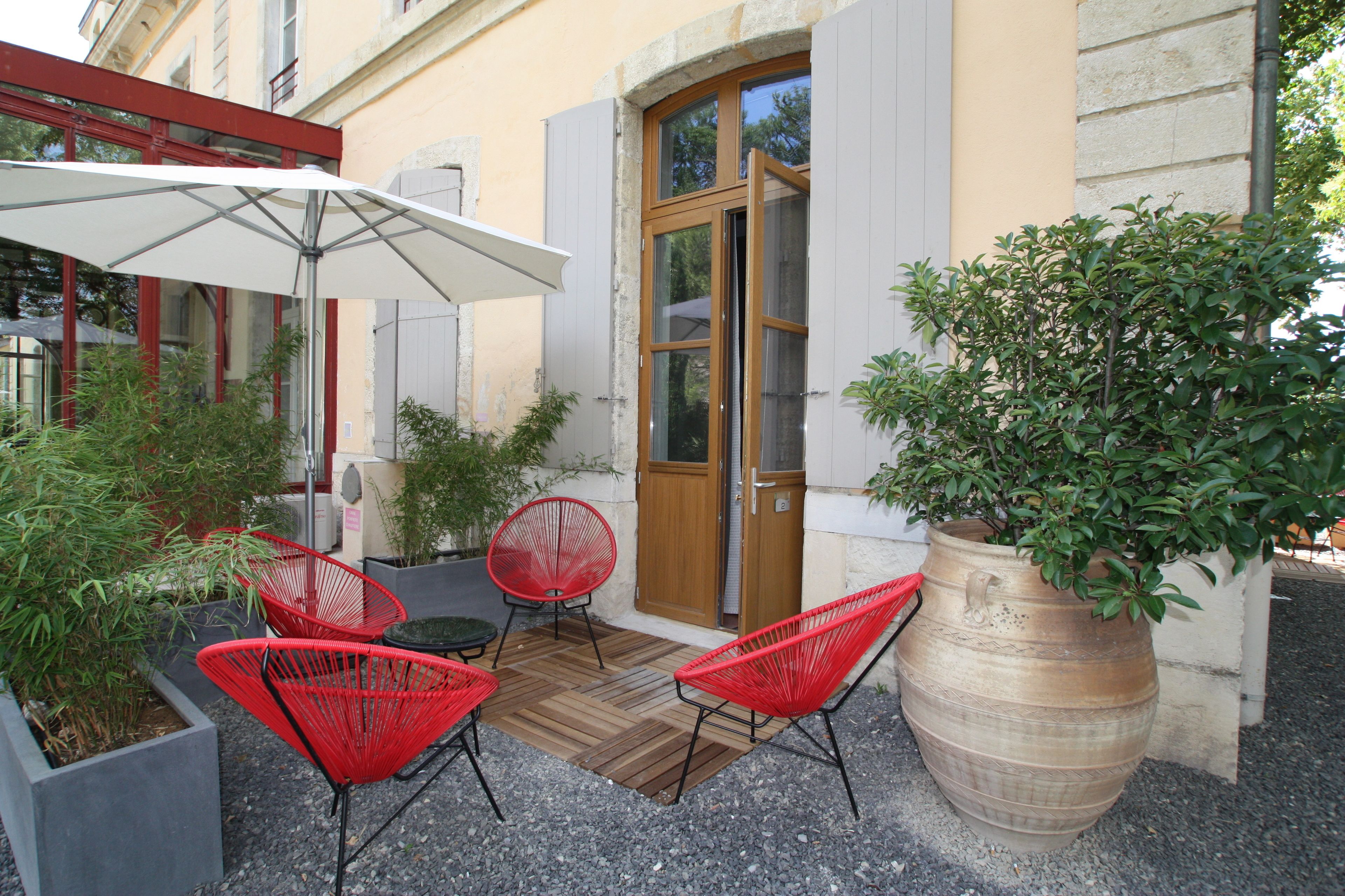 An outdoor seating area with red chairs and a white umbrella, a large potted plant, and a wooden door.