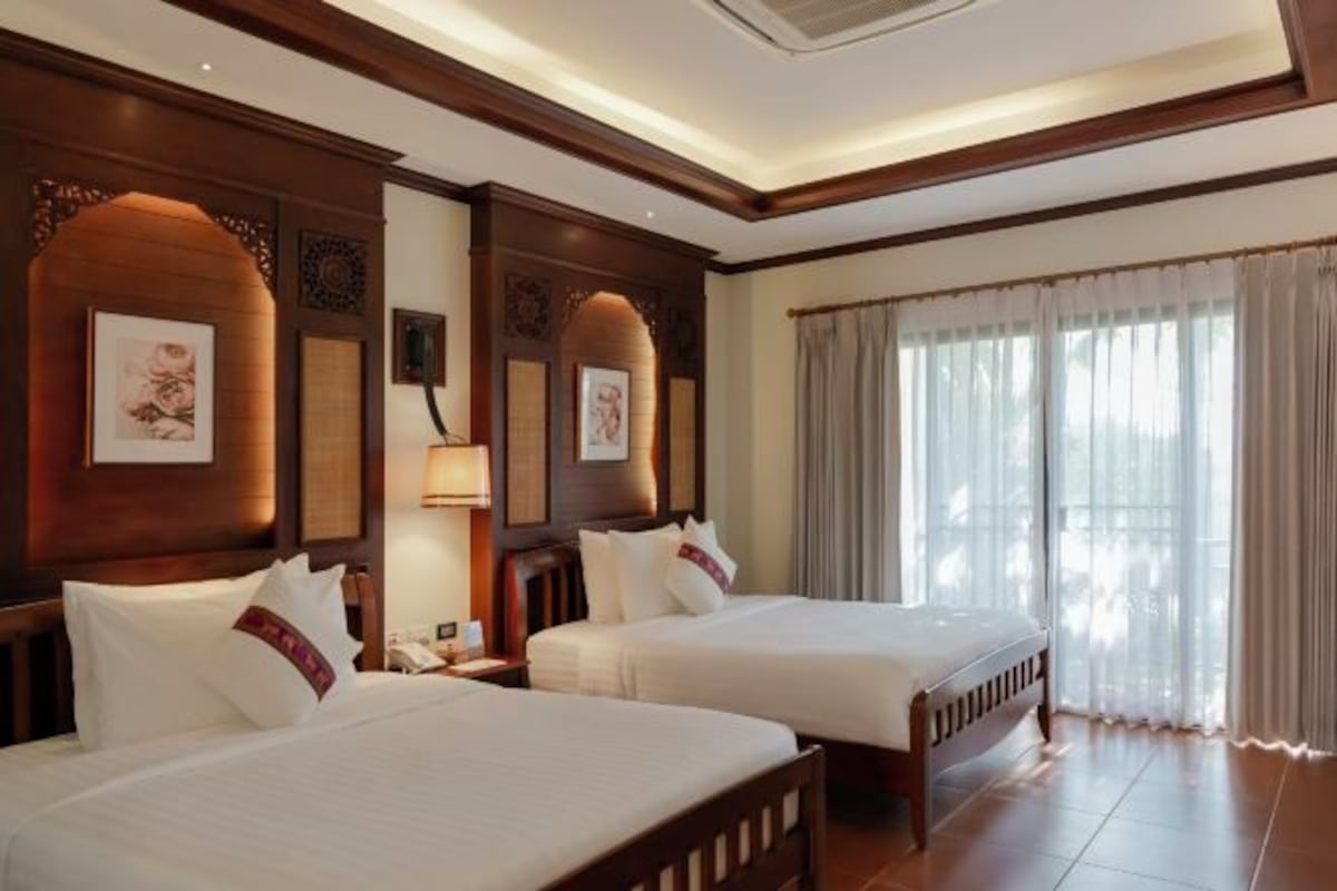 A hotel room with two beds, wooden headboards, a large window with curtains, and wall-mounted artwork.