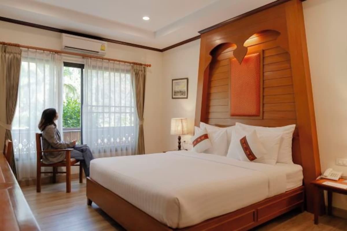 A hotel room with a large bed, a desk, and a chair. There is a window with curtains, a lamp on the desk, and a phone on the nightstand.