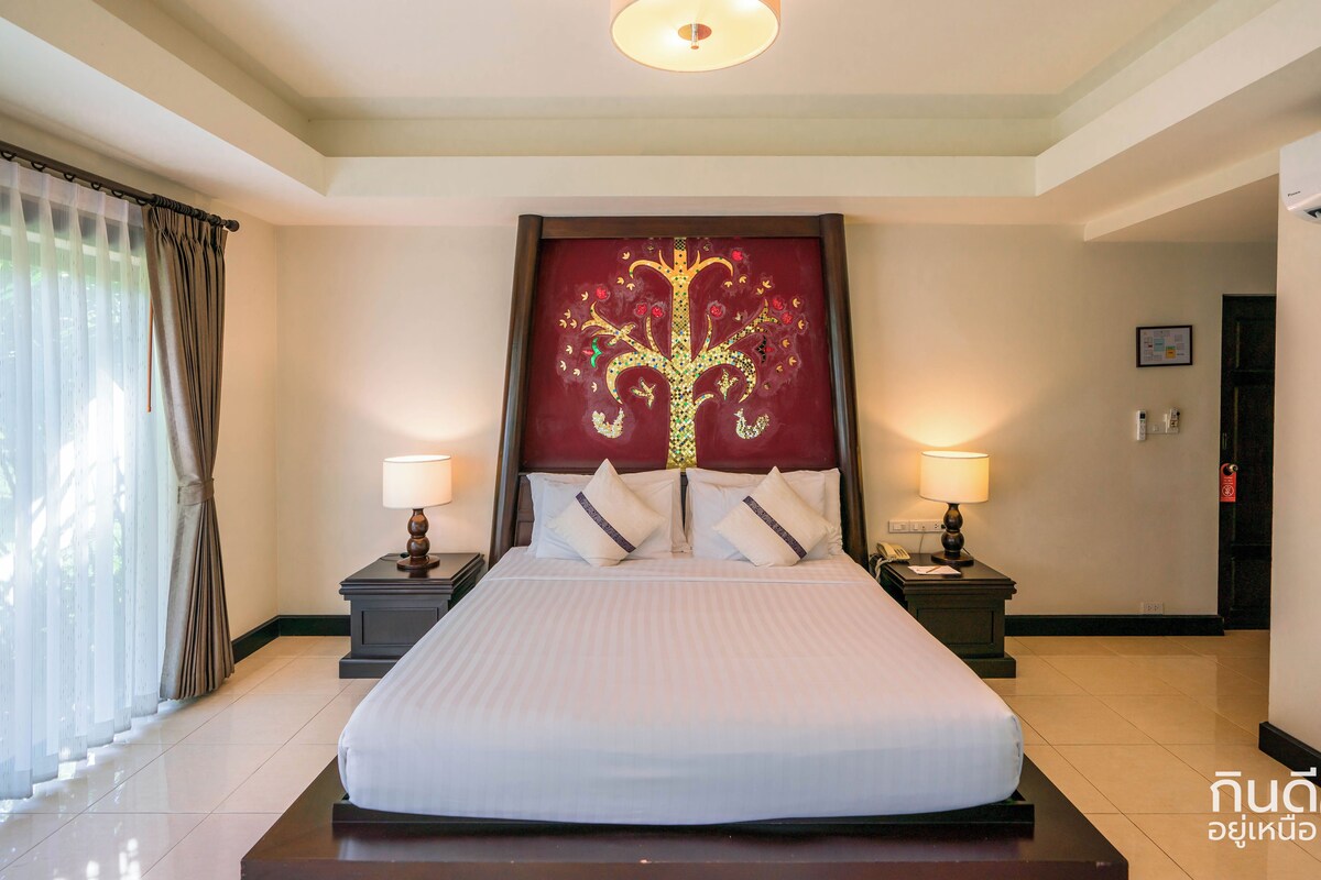 A hotel room with a large bed, two bedside tables with lamps, a decorative headboard, and a view of the outside through sheer curtains.