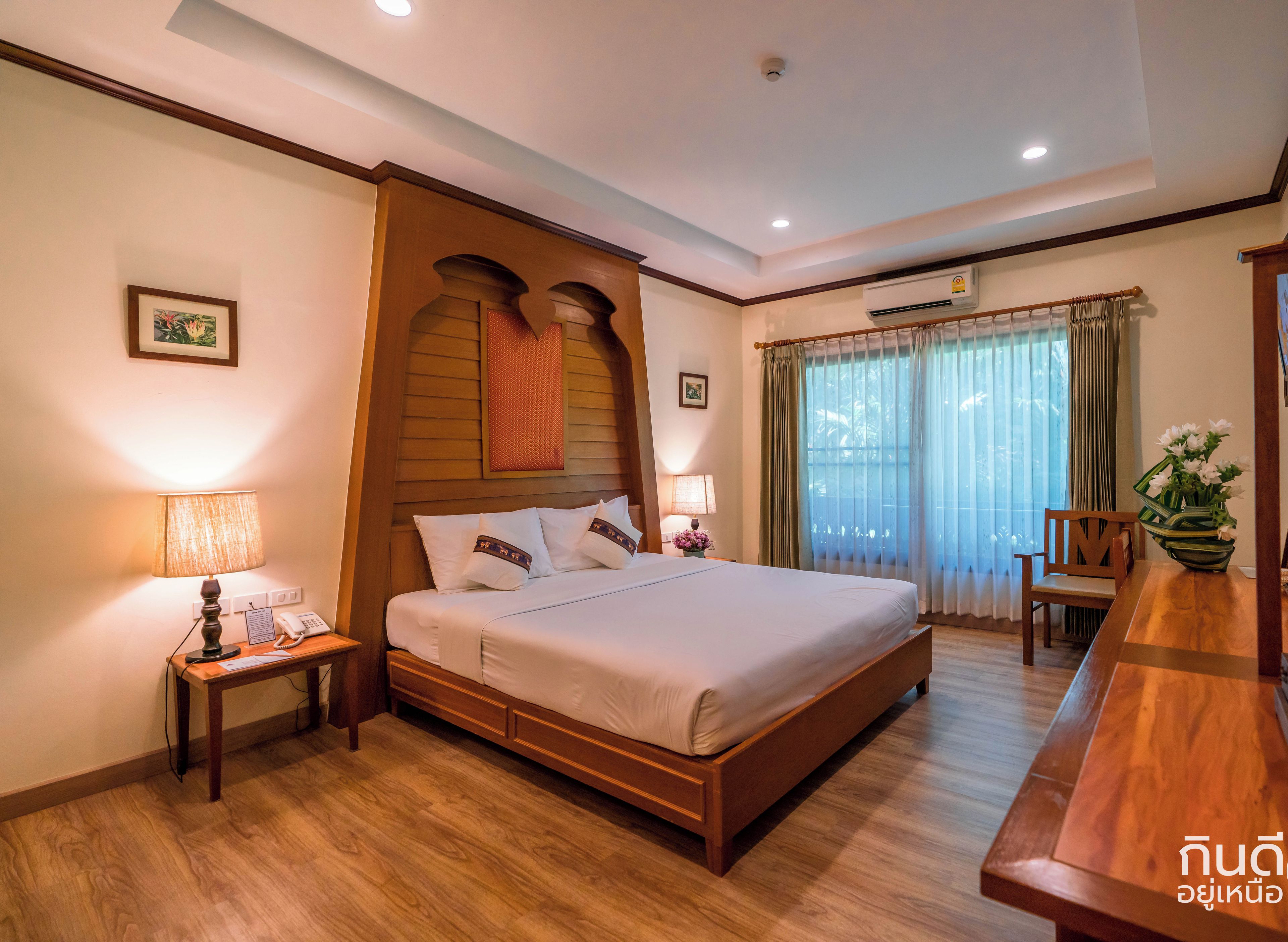 A hotel room with a wooden bed, bedside tables with lamps, a desk with a chair, and a large window with curtains.