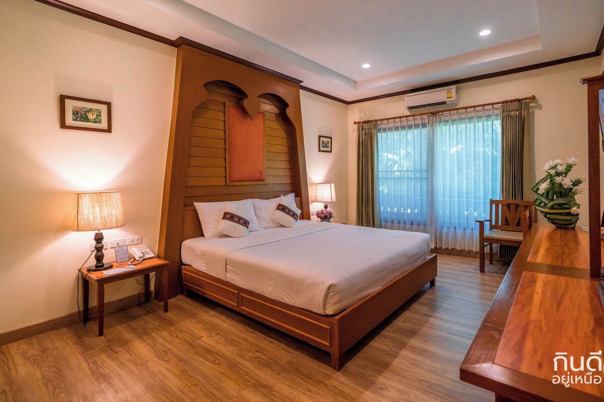 A hotel room with a wooden bed, bedside tables with lamps, a desk with a chair, and a large window with curtains.