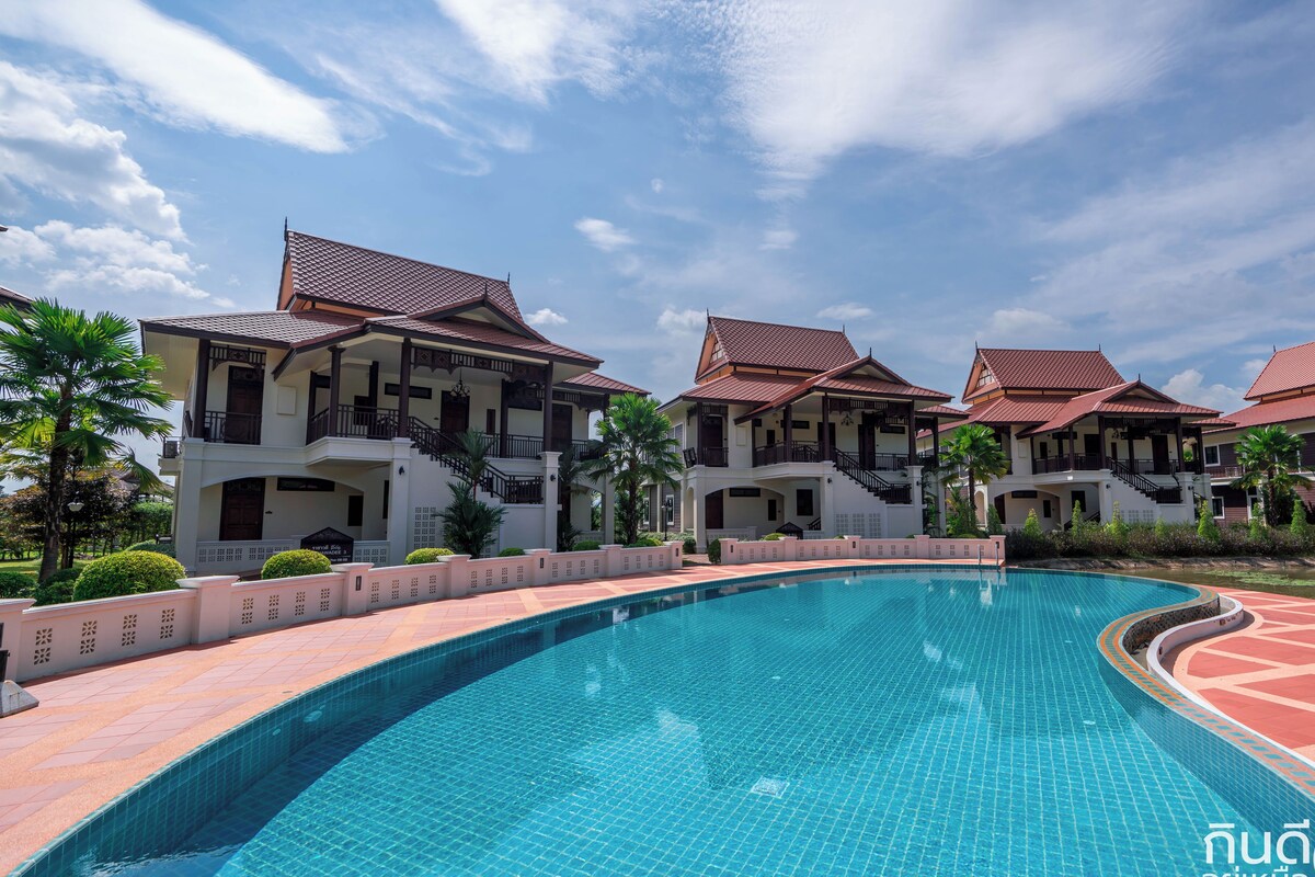 A swimming pool with a curved edge, surrounded by a tiled deck and a row of traditional-style buildings with red roofs and balconies.