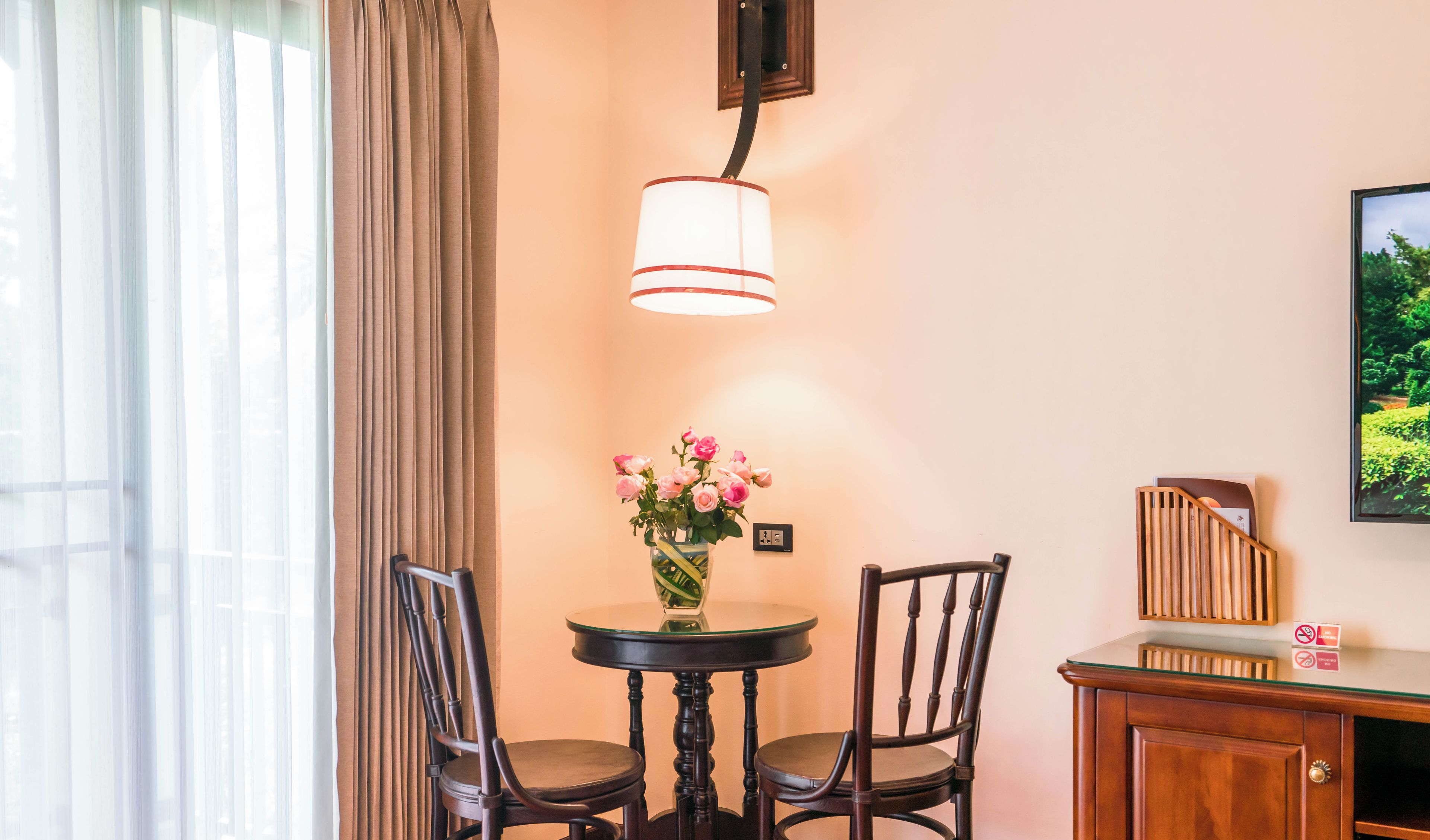 Una habitación de hotel con una mesa redonda, dos sillas, un florero con flores, una lámpara, un armario y un televisor en la pared.