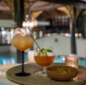 Two cocktails on a table with a candle and a bowl of snacks.