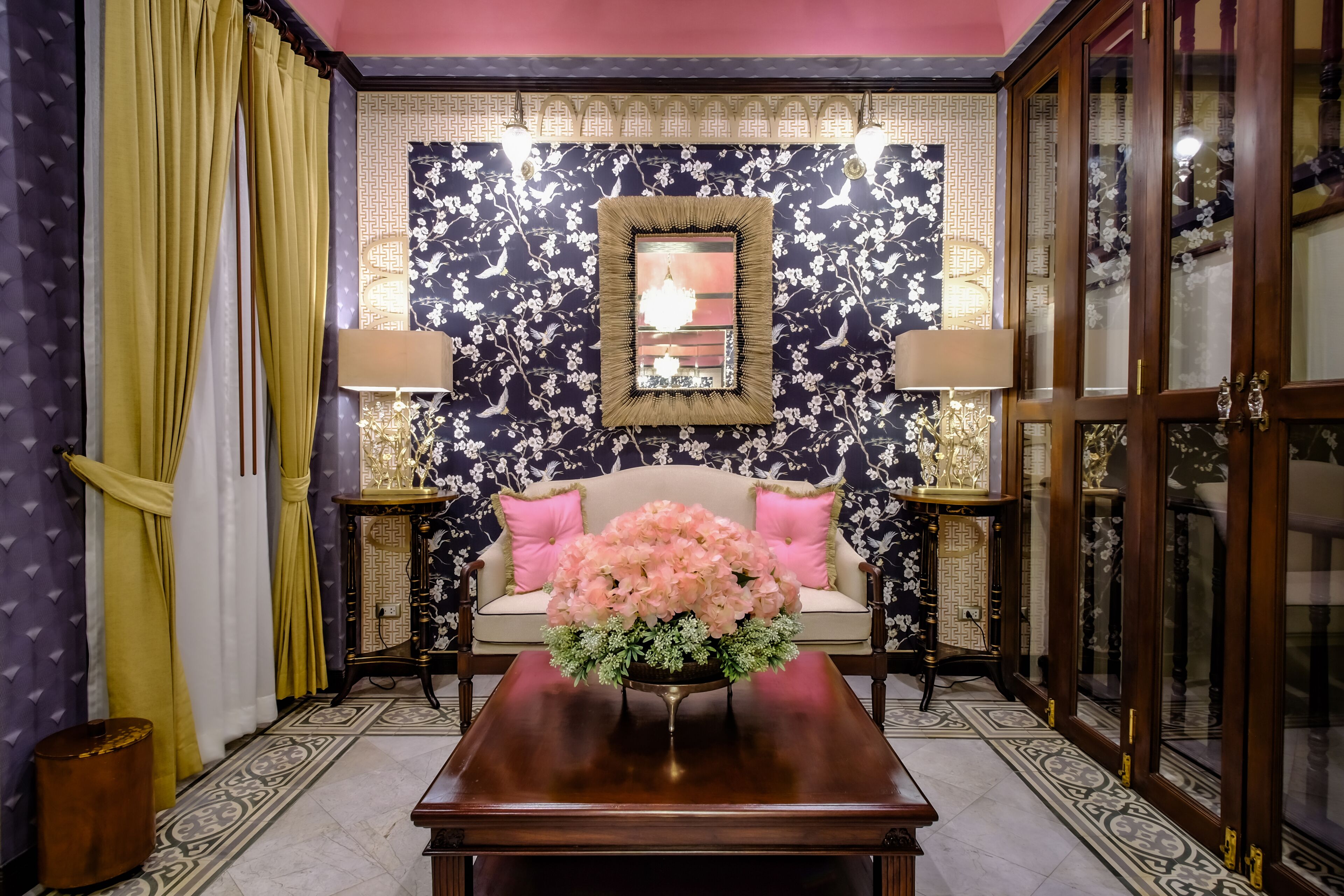 A room with a sofa, a coffee table with a floral arrangement, a mirror, and patterned wallpaper.