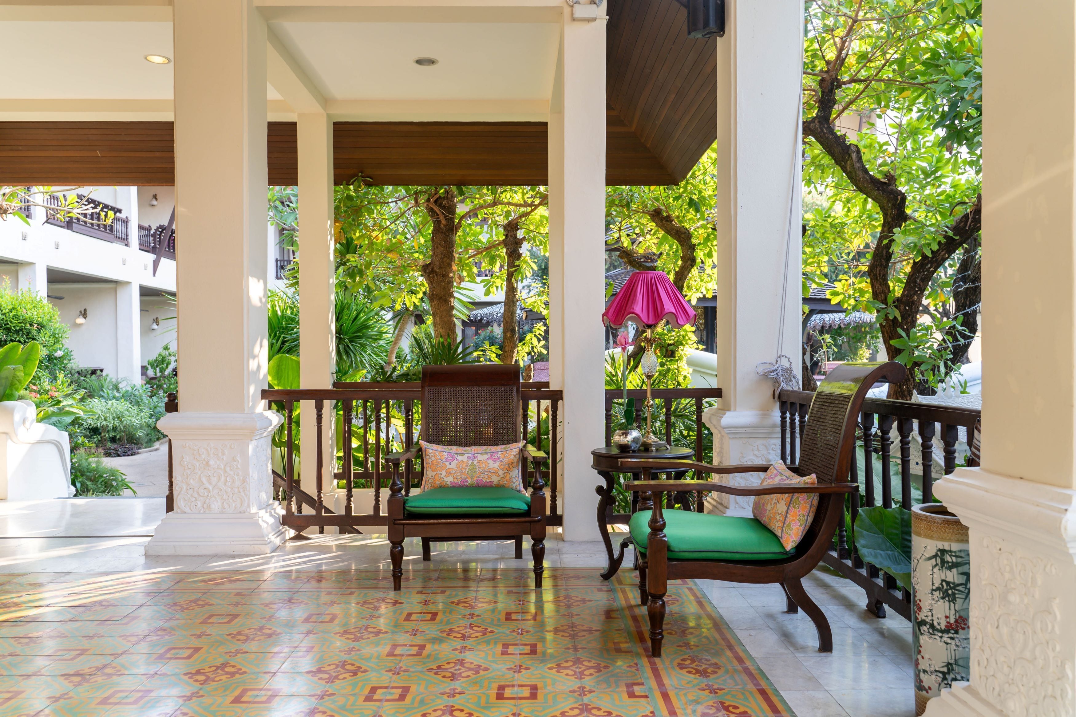 A covered outdoor seating area with wooden chairs and a patterned floor, surrounded by greenery and a view of a building in the background.