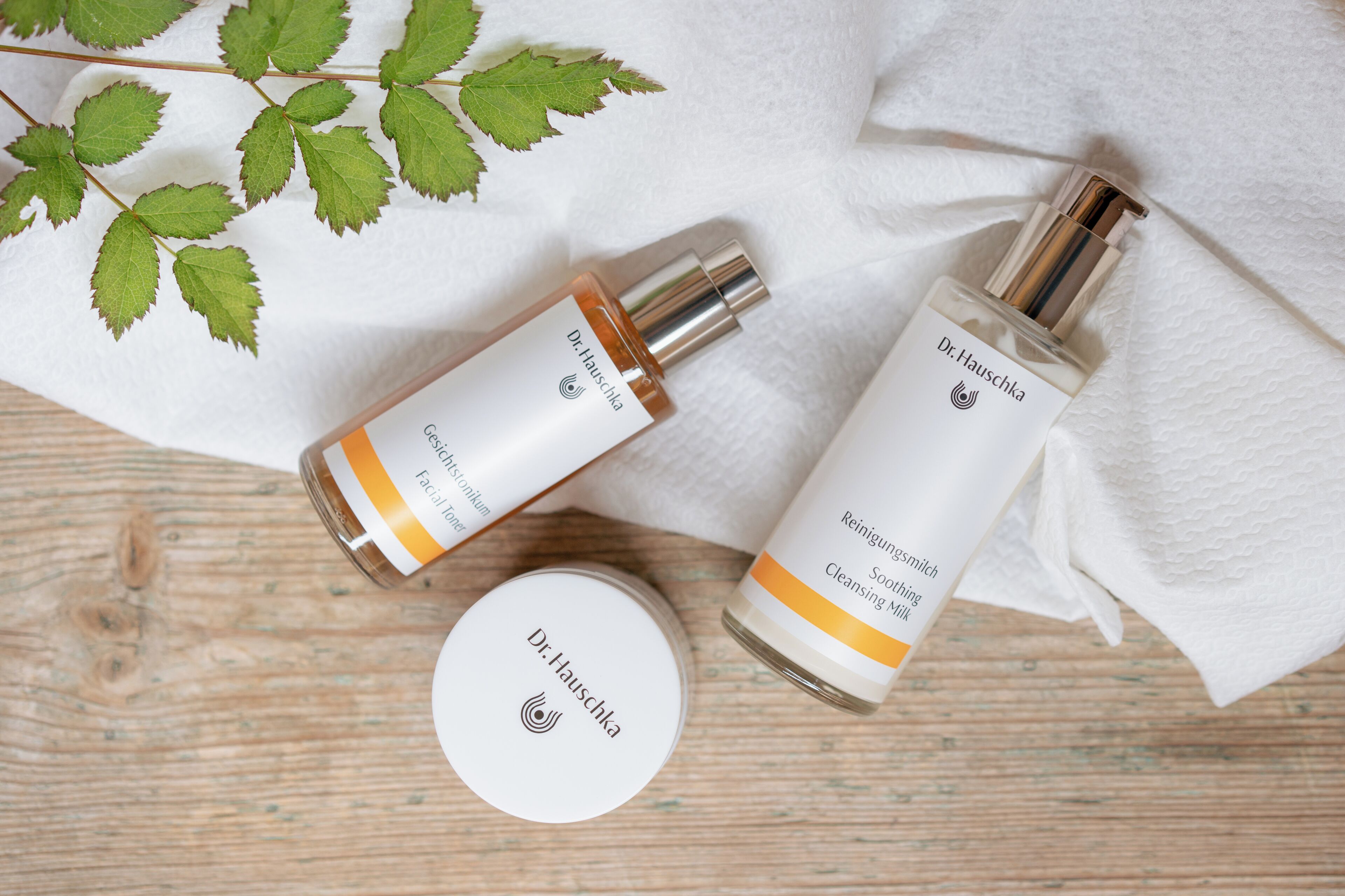 Two bottles of Dr. Hauschka skincare products and a container on a wooden surface with a white cloth.