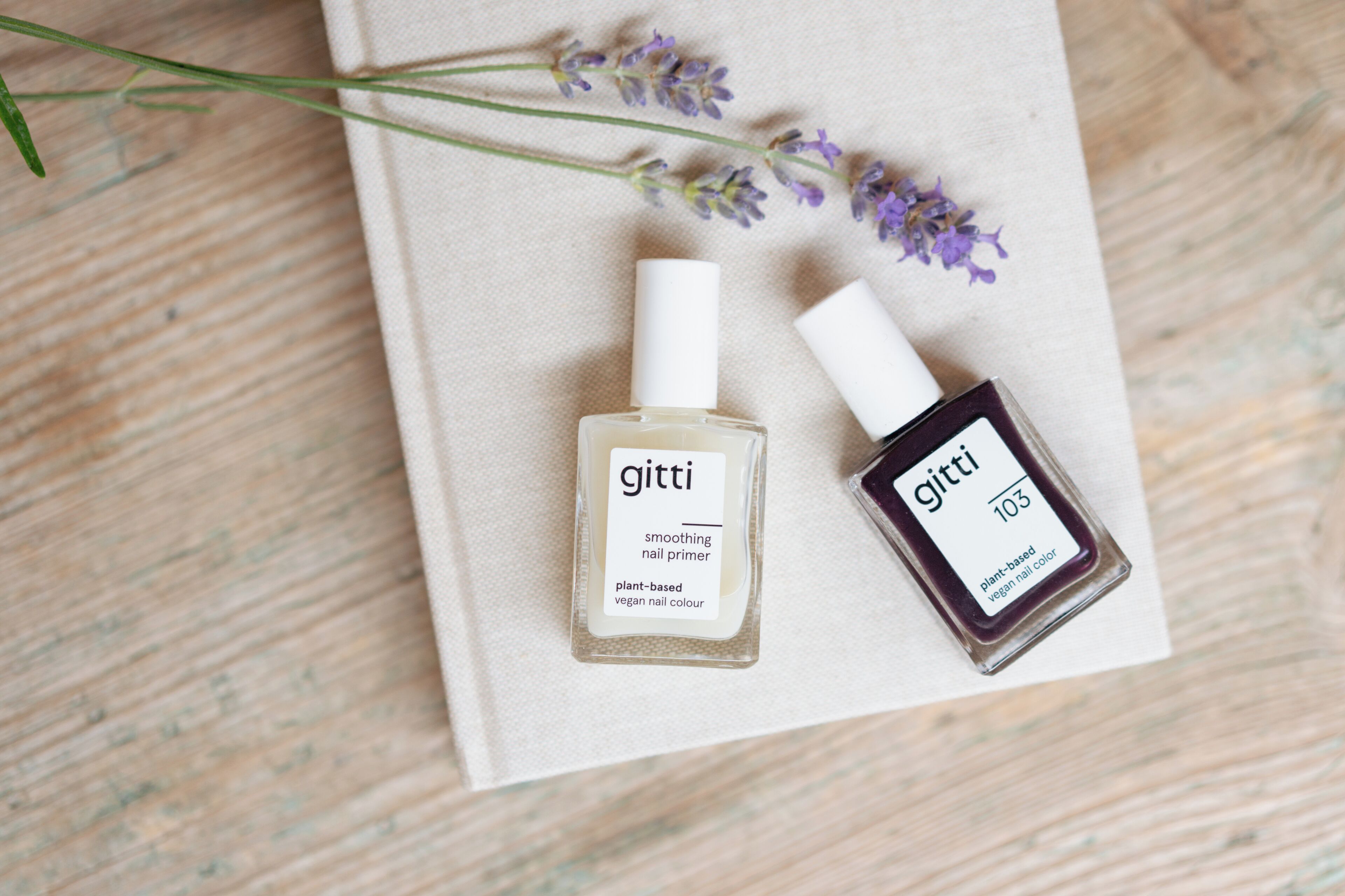 Two bottles of Gitti nail primer on a beige fabric surface with lavender flowers.