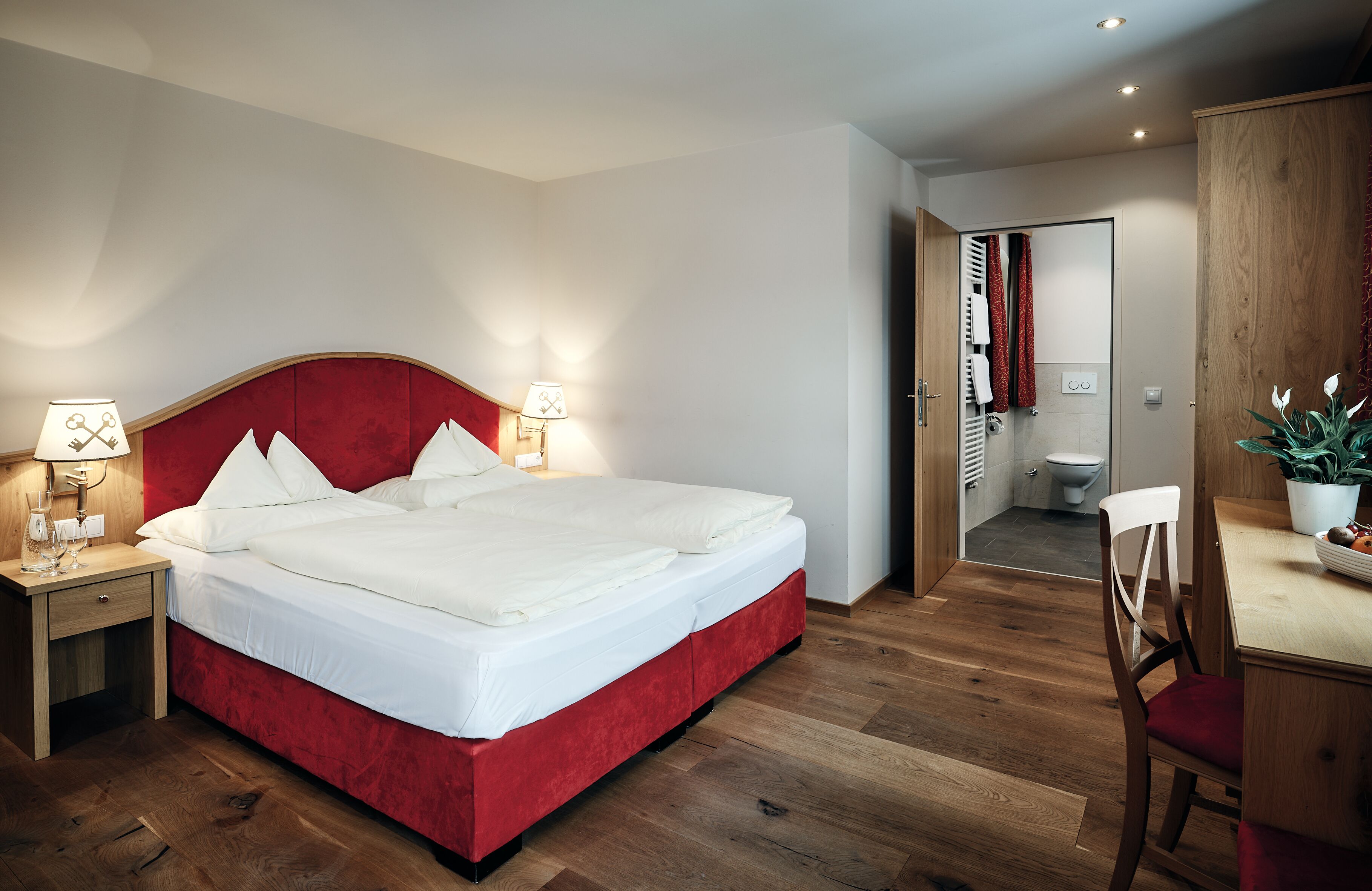 A modern bedroom with a red upholstered bed, bedside lamps, a wooden nightstand, a wooden desk with a chair, and a potted plant.
