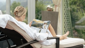 Una persona leyendo una revista en una habitación soleada con vistas a zonas verdes y montañas.