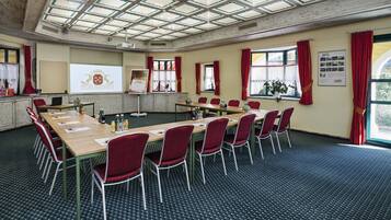 A conference room with a long table, red chairs, a projector screen, and a large window with curtains.