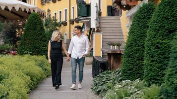 A couple walking on a paved path in a garden area with a yellow building in the background.