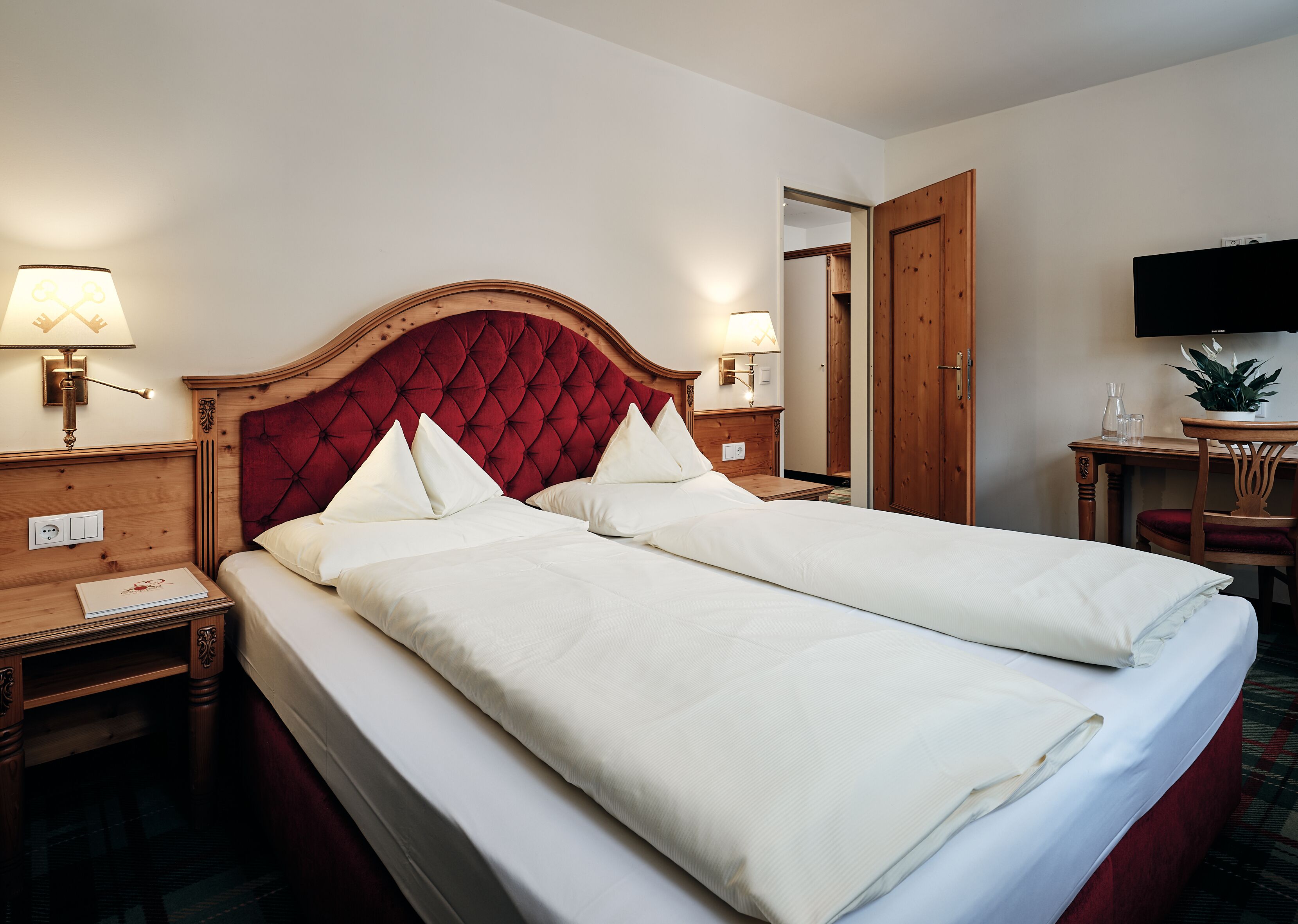 Ein Hotelzimmer mit einem großen Bett, zwei Nachttischen, einem hölzernen Kopfteil, einem Fernseher und einem kleinen Tisch mit einer Pflanze.