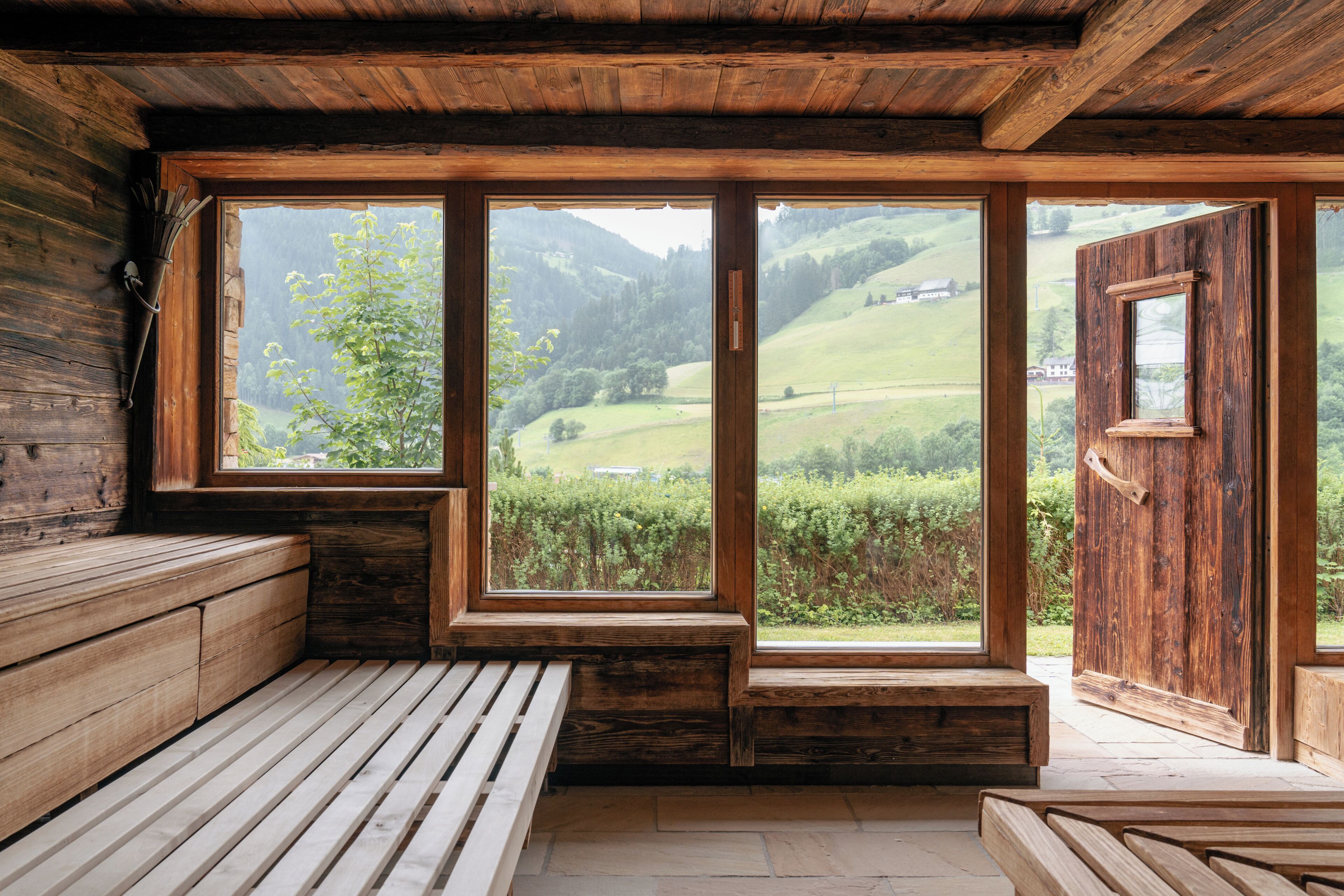 A wooden cabin with large windows offering a view of a green valley.