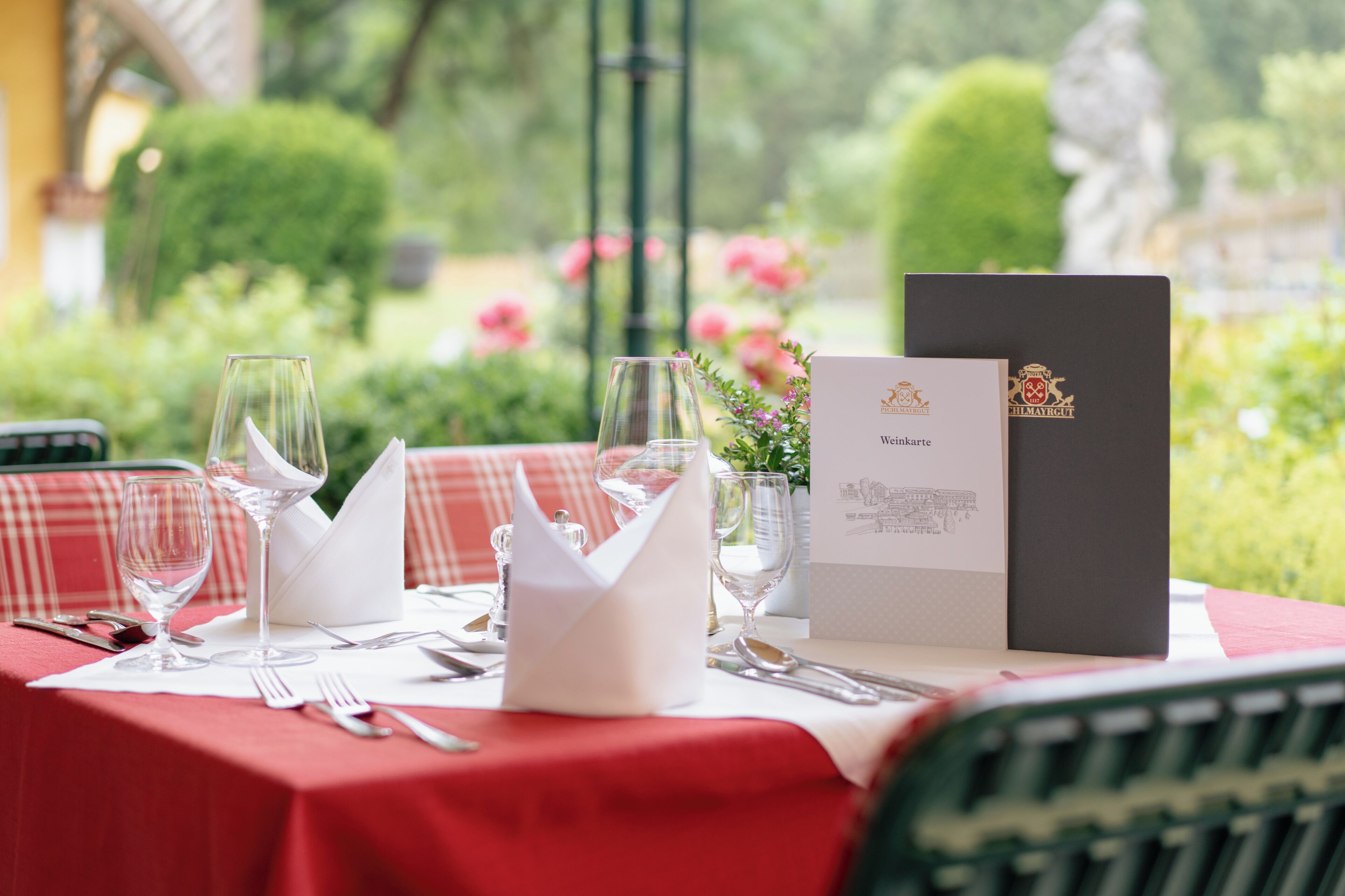 A table set for dining outdoors with a red tablecloth, wine glasses, cutlery, and a menu.