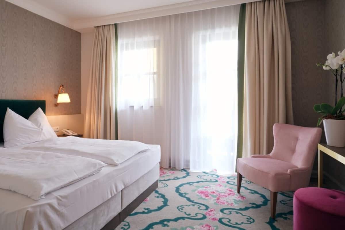 A hotel room with a bed, a chair, a nightstand, a lamp, and a window with curtains.