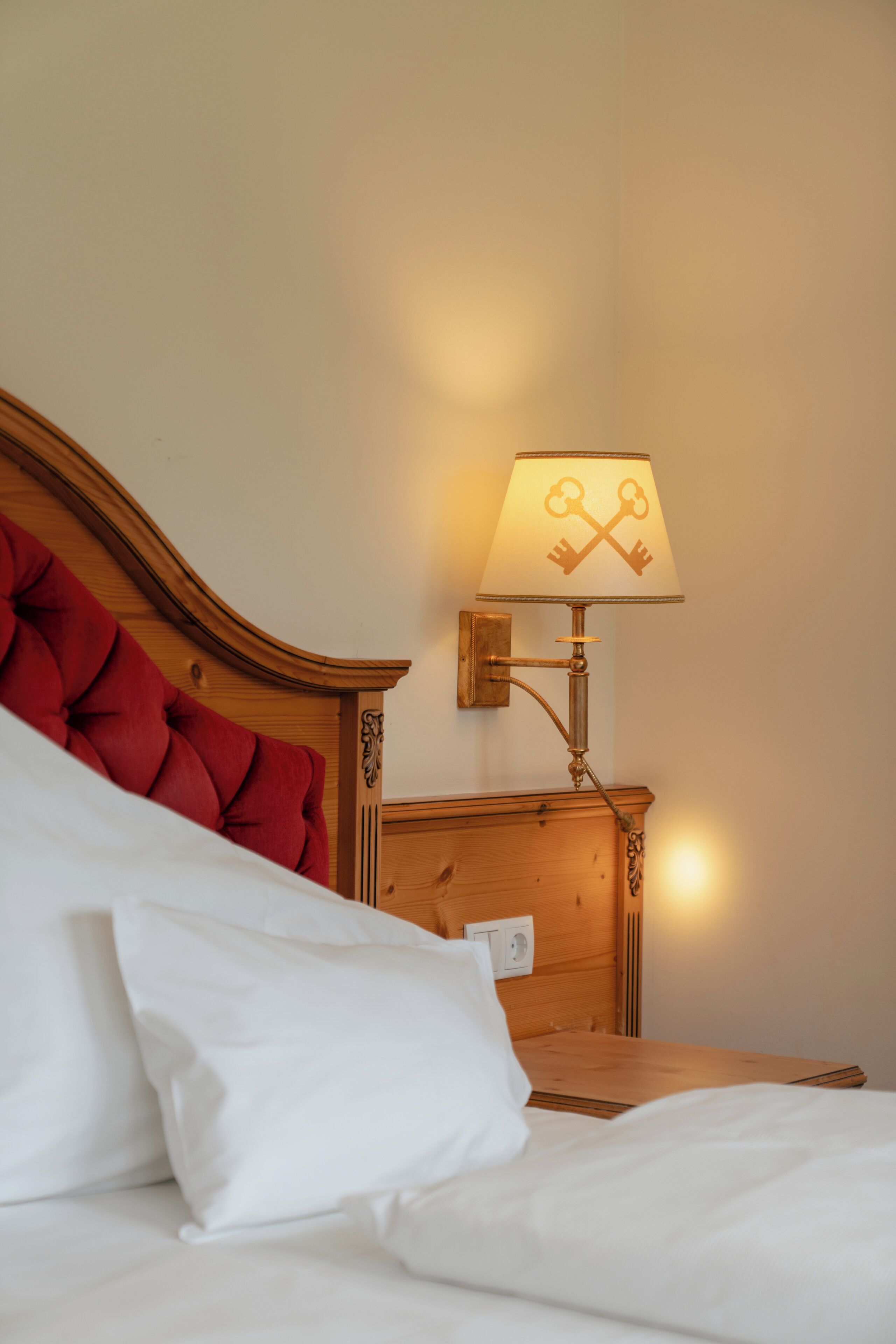 A neatly made bed with white linens and a red tufted headboard. A wall-mounted lamp with a key design is visible.